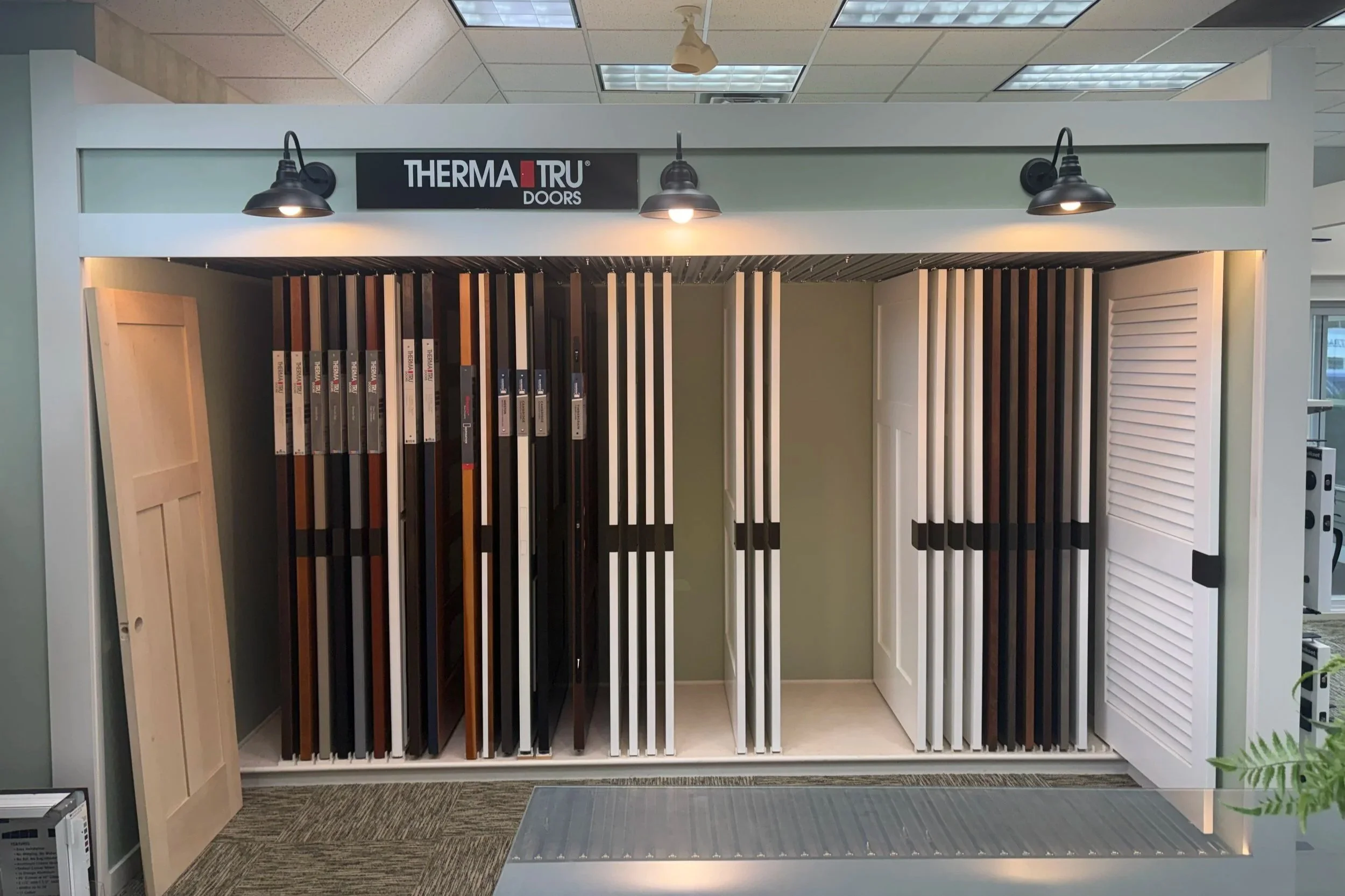 Showroom display of various door styles and colors, with the sign 'Therma-Tru Doors' at the top. The display includes sample door panels and slats, and the area is lit by ceiling lights.
