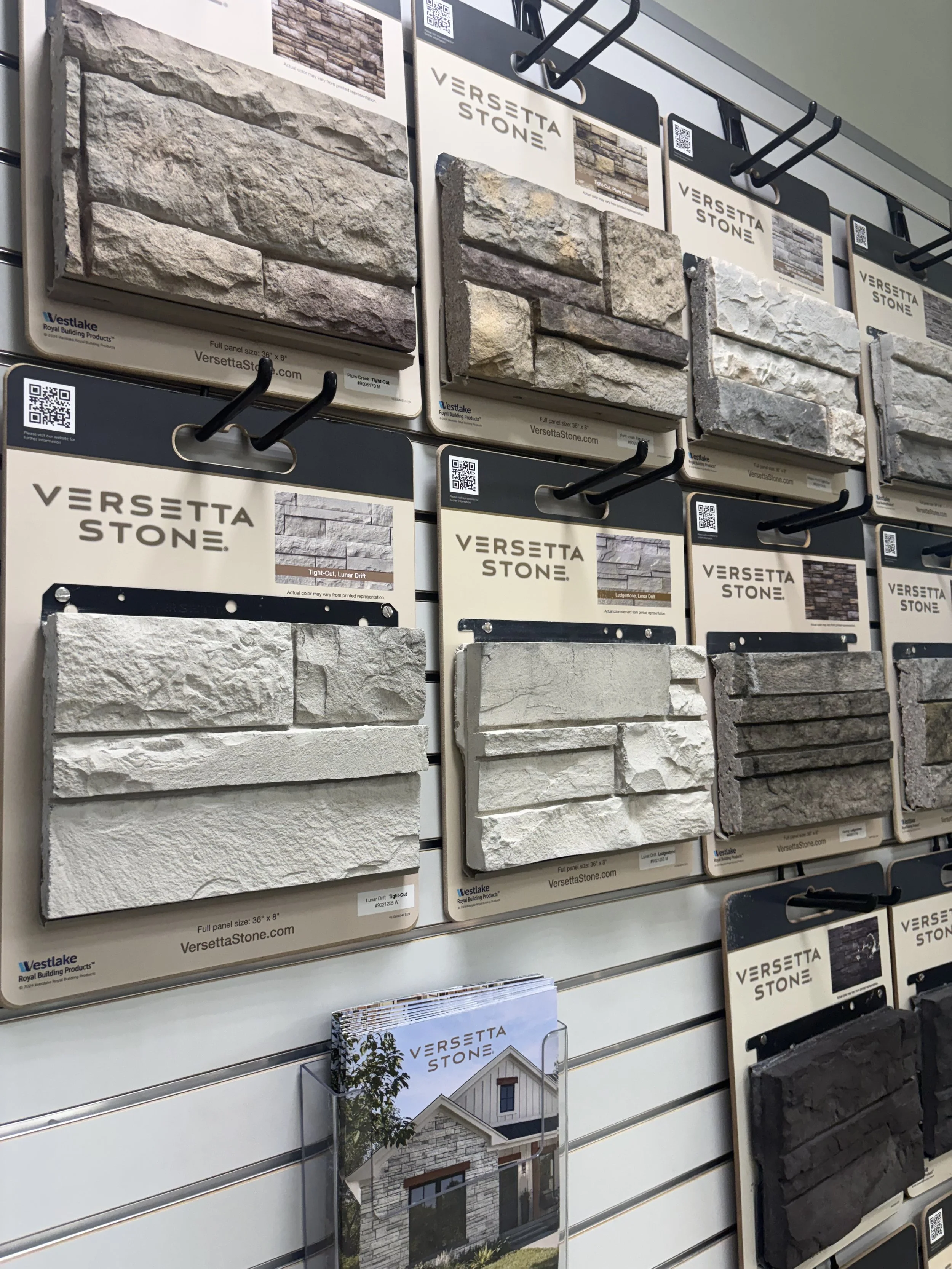 Display of stone veneer samples on a store wall, with sample panels labeled 'Versetta Stone' in various colors and textures, arranged on hooks.