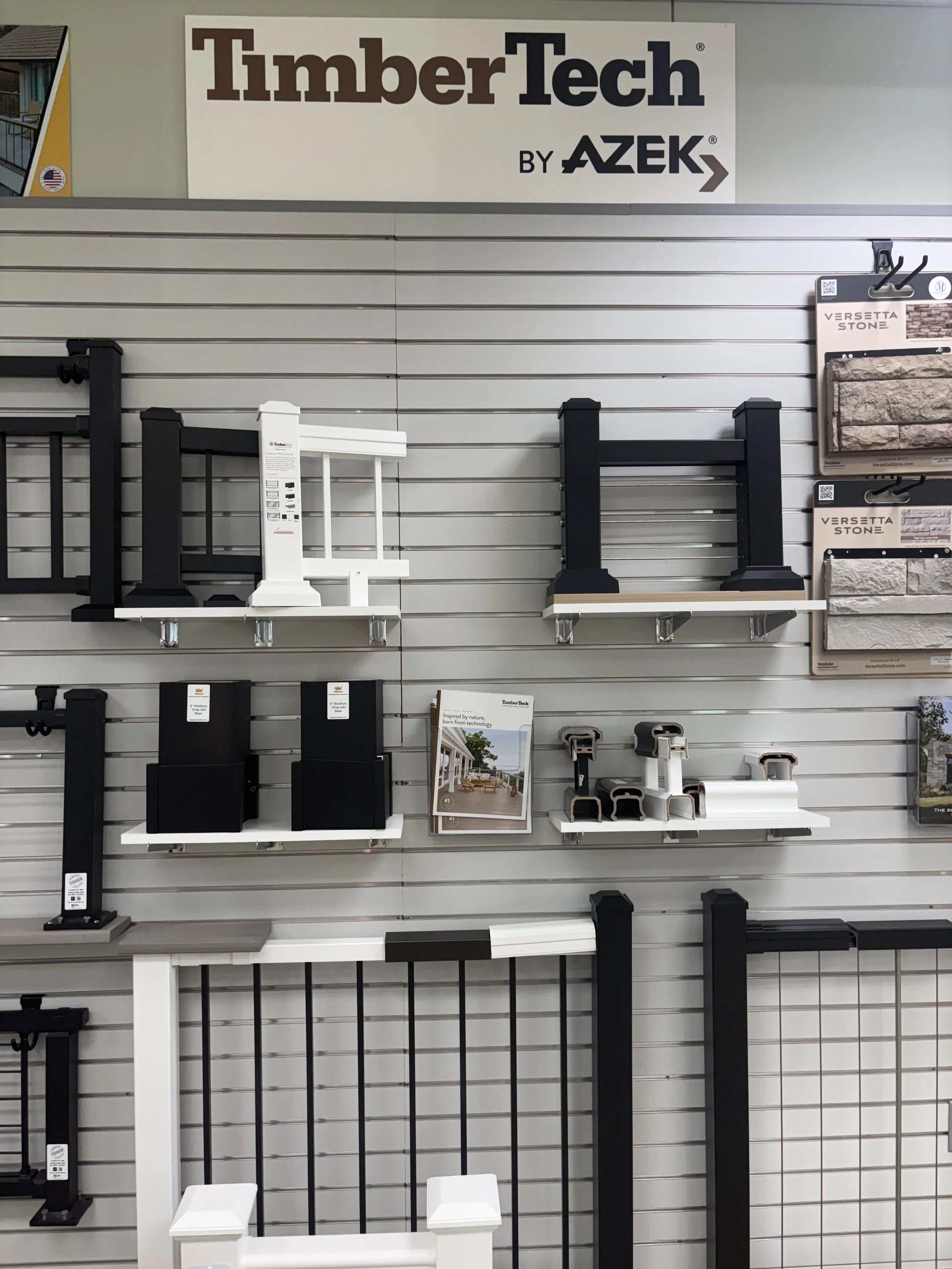 Display of building materials and hardware on a store wall, including black and white trim and siding samples, with a 'TimberTech by AZEK' sign at the top.