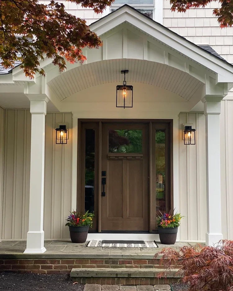 Exterior view of a house front door with a wooden door, two wall-mounted lantern-style lights, a pendant ceiling light, and potted plants with colorful flowers on either side of the door.