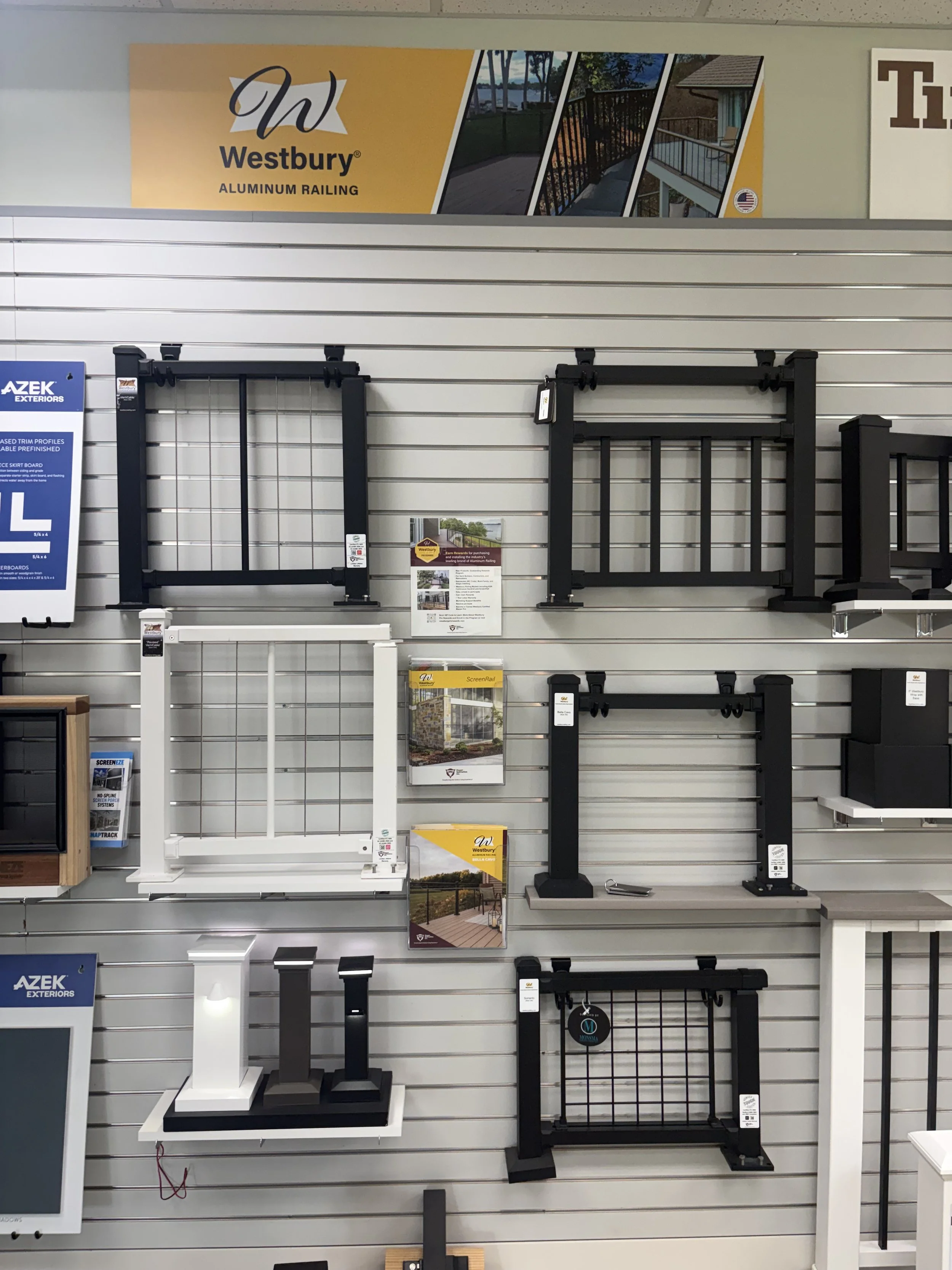 Display of various aluminum railing styles and colors on a showroom wall for Westbury brand, including black and white options, with informational brochures and signage.