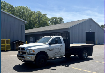 Woodwork Specialties' white flatbed truck parked outside a our big gray barn with a big overhead garage door and yellow storage bins nearby.