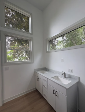 A small white laundry or utility sink with cabinet storage underneath, situated in a bright room with three large windows showing greenery outside.