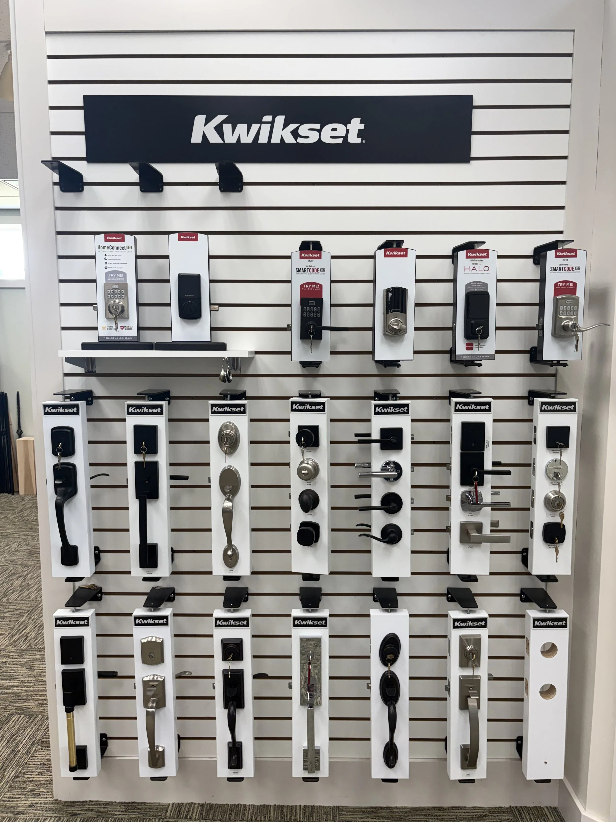 Display of various black, silver, and gold door handles, locks, and related hardware on a white slatwall.