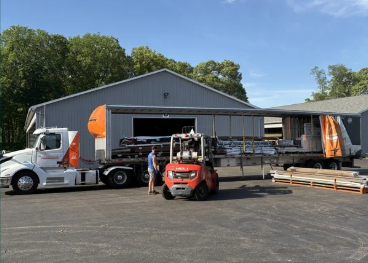 Workers unloading building supplies and equipment onto trucks at an industrial building with trees in the background.