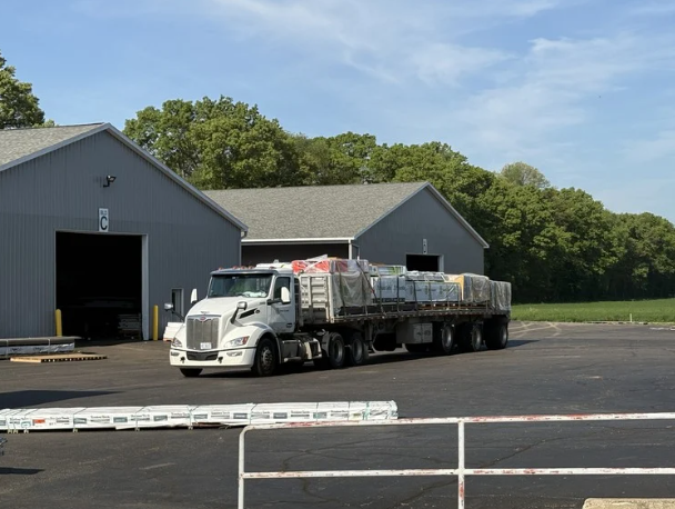 A large white delivery semi-truck parked in front of Woodwork Specialties' warehouse with an open garage door, carrying load of lumber and building supplies.