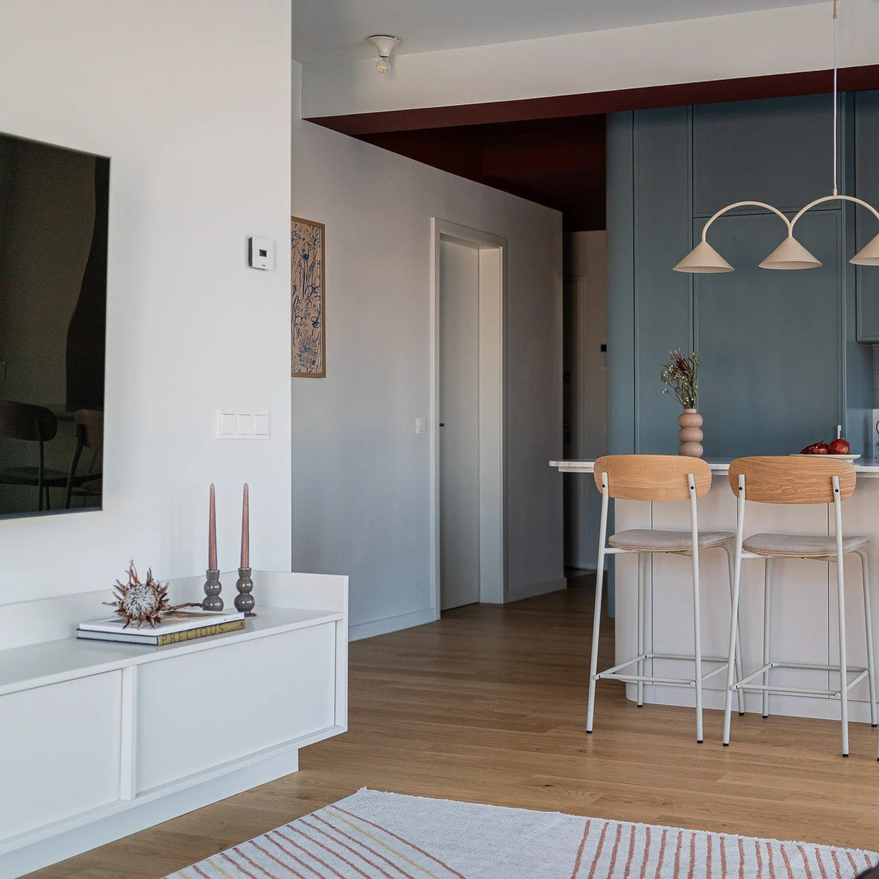 Modern kitchen with blue cabinetry, marble island, pendant lights, large window with sheer curtains, and dining area with wooden chairs and a sofa.