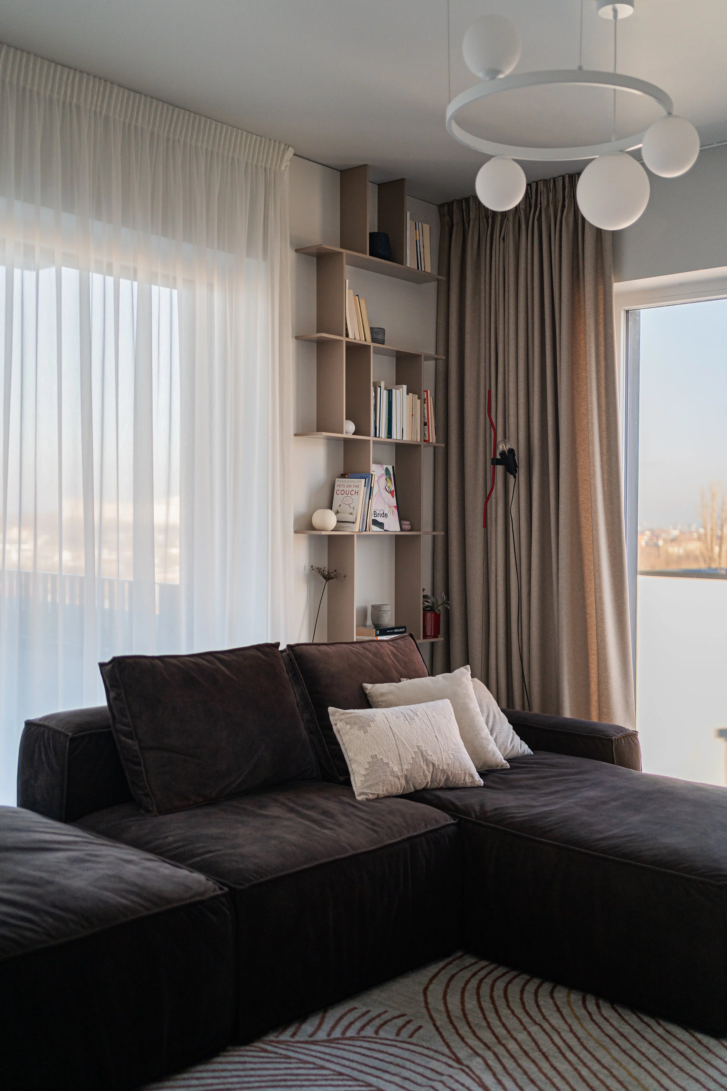 Modern living room with beige sectional sofa, round coffee tables, TV on white wall, large windows with curtains, ceiling light fixture, indoor plant, and neutral decor.