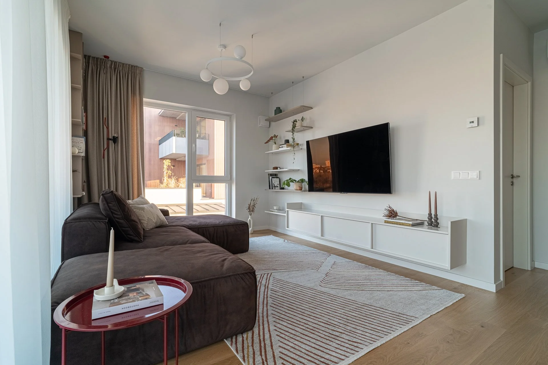 Modern living room with beige sectional sofa, round coffee tables, TV on white wall, large windows with curtains, ceiling light fixture, indoor plant, and neutral decor.