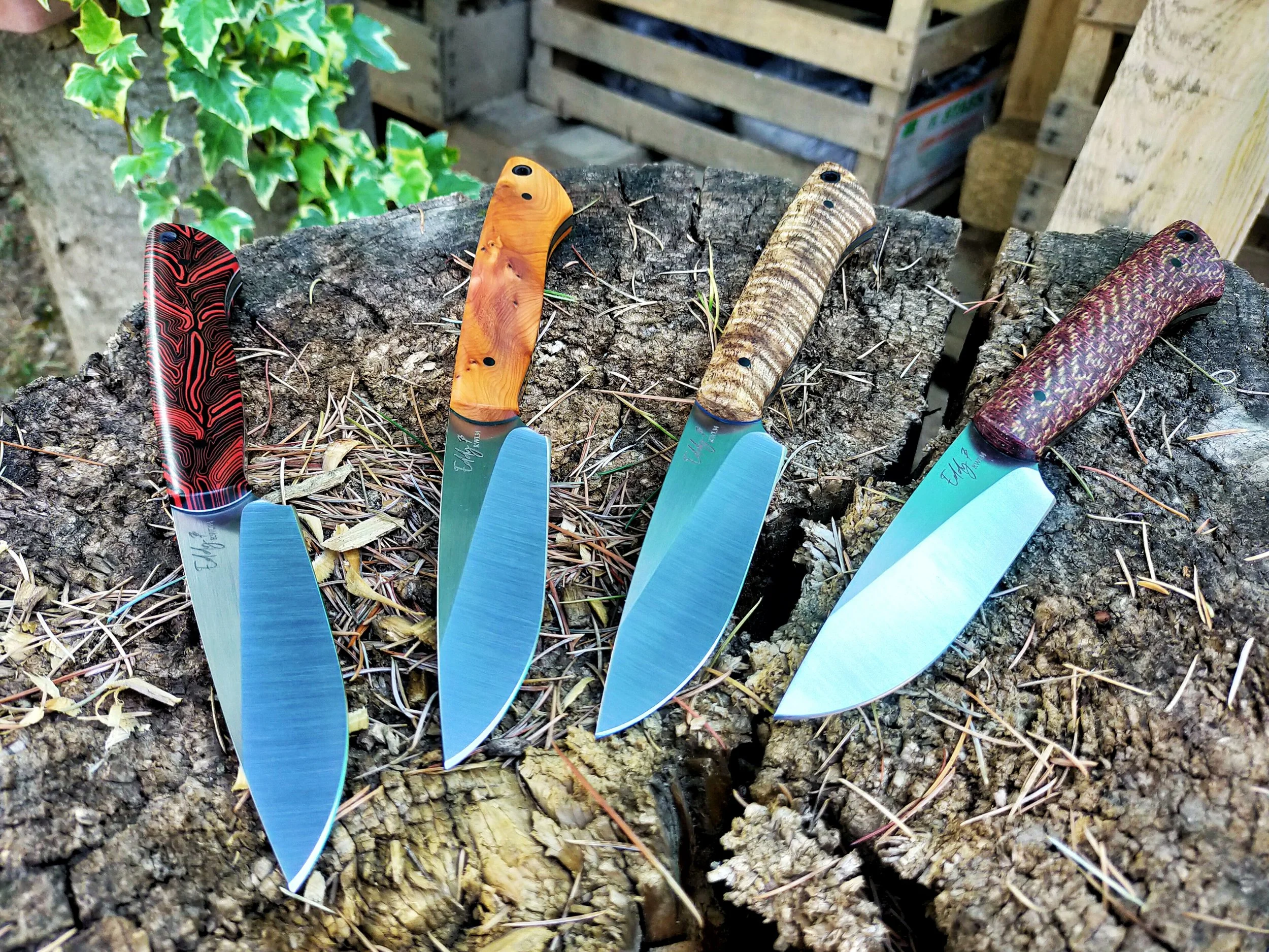 Five colorful knives with patterned handles arranged on a tree stump outdoors.