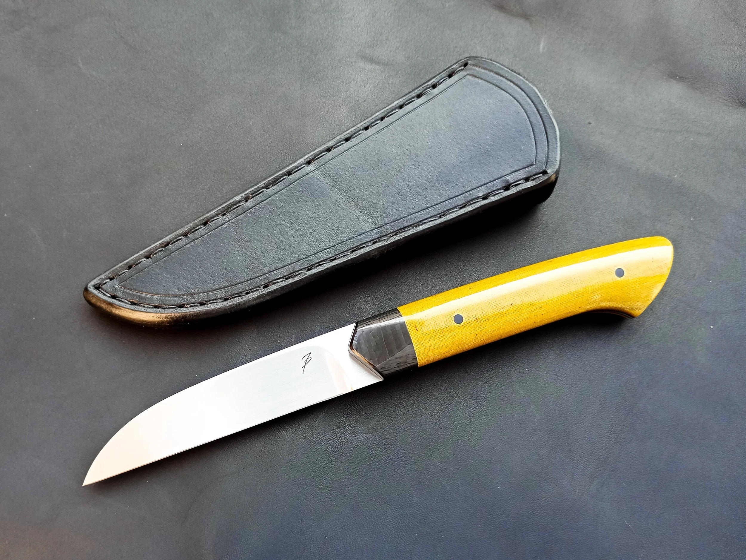 A EDC knife with a yellow micarta handle next to a black leather sheath, both placed on a black surface.