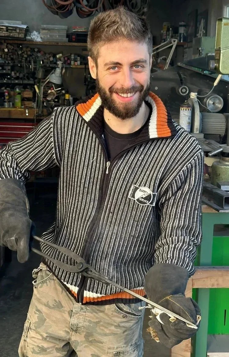 A smiling man with a beard and short hair working in a workshop, holding a metal tool.