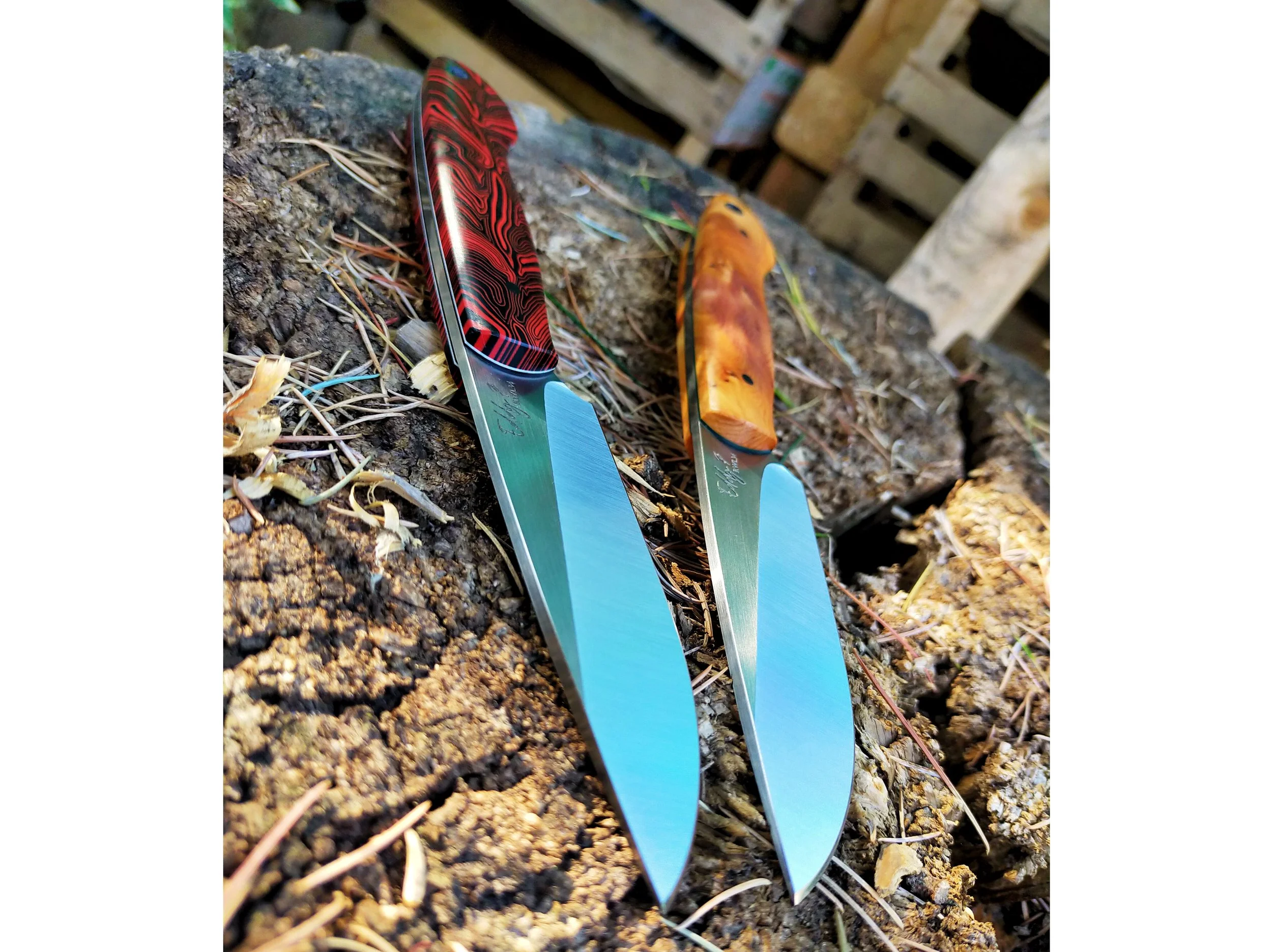 Two knives with colorful handles and stainless steel blades resting on soil and small sticks outside.