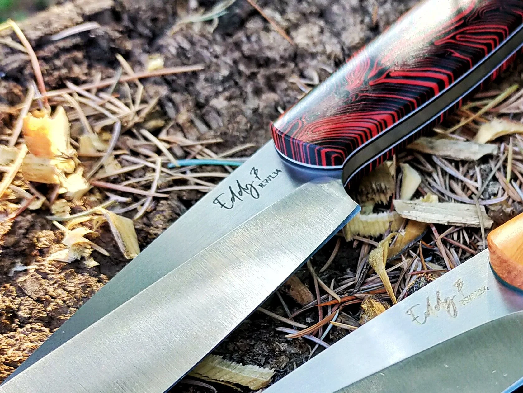 Two hunting knives with polished blades and black and red patterned handles lying on dirt and dry grass.
