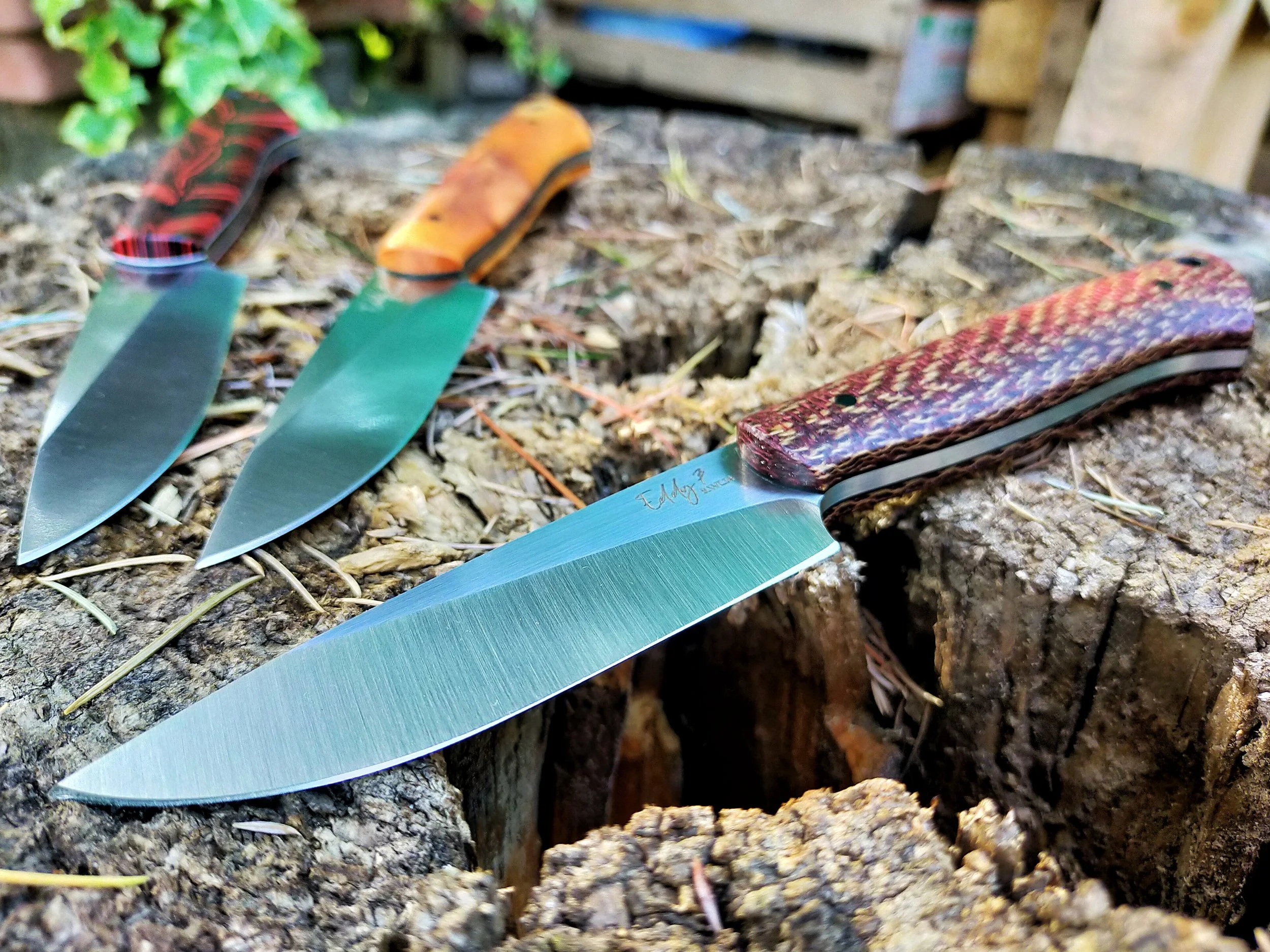 Three hunting knives with colorful handles placed on a tree stump outdoors.
