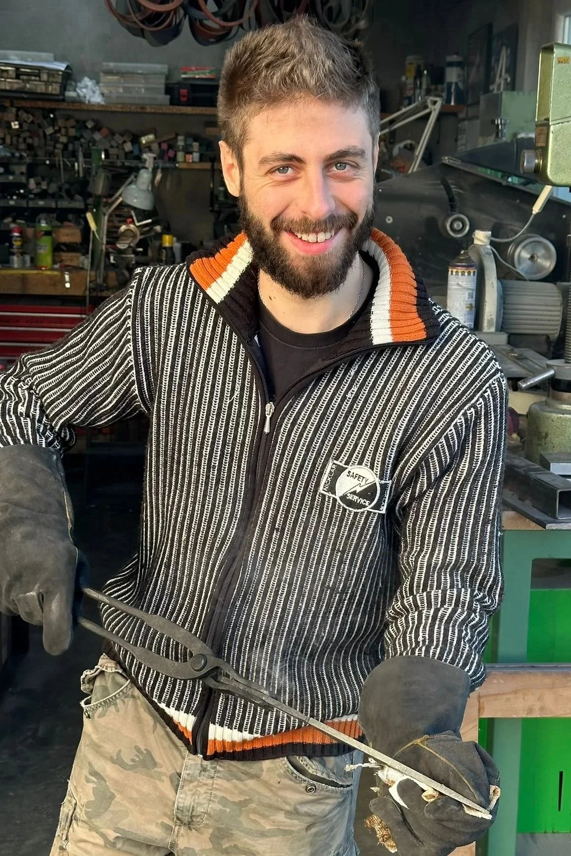 A smiling man with a beard and short hair working in a workshop, holding a metal tool.