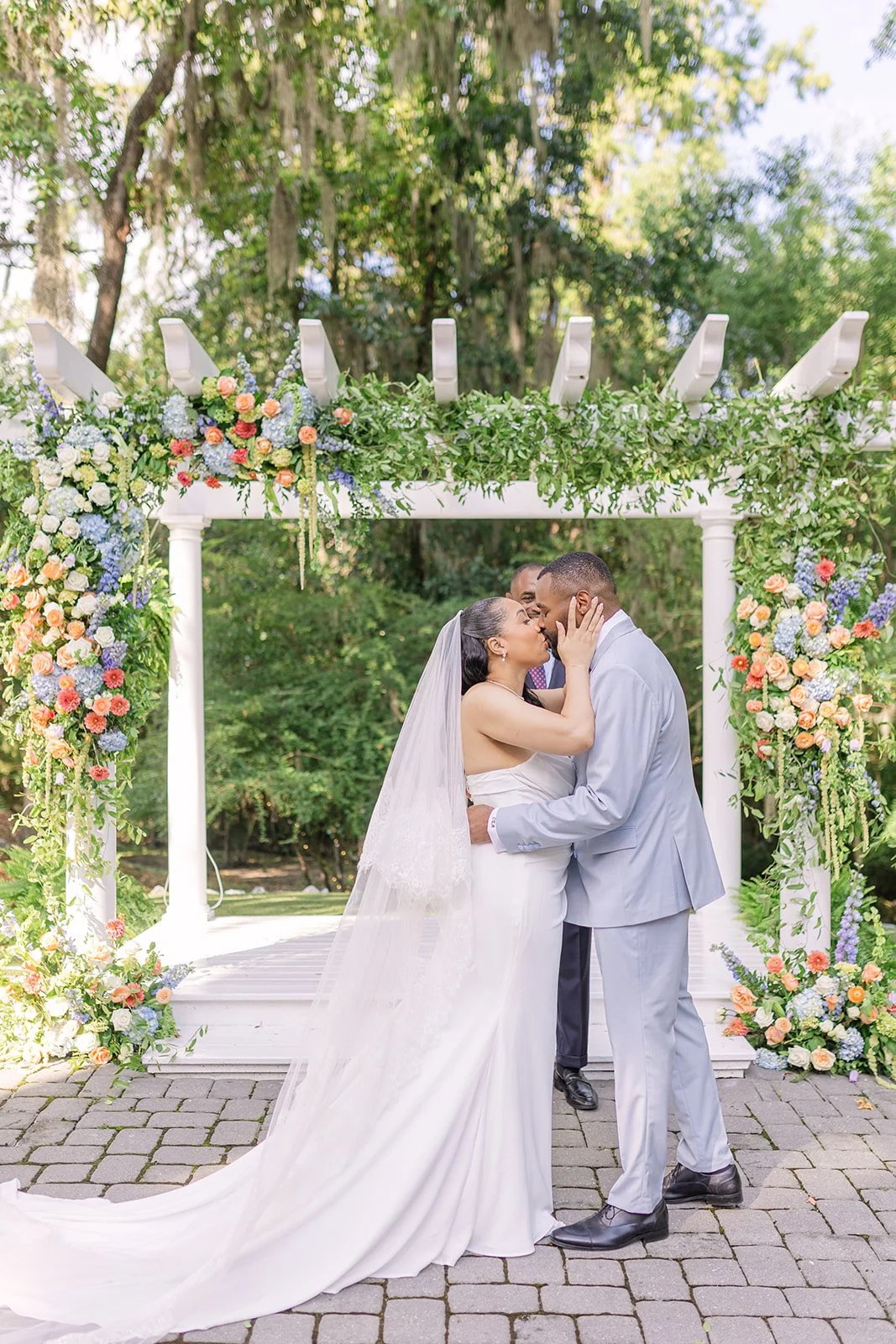 Bride and groom kissing beneath colorful floral ceremony arbor at The Mackey House wedding venue in Savannah Georgia featuring lush garden wedding flowers and outdoor Southern ceremony setting.