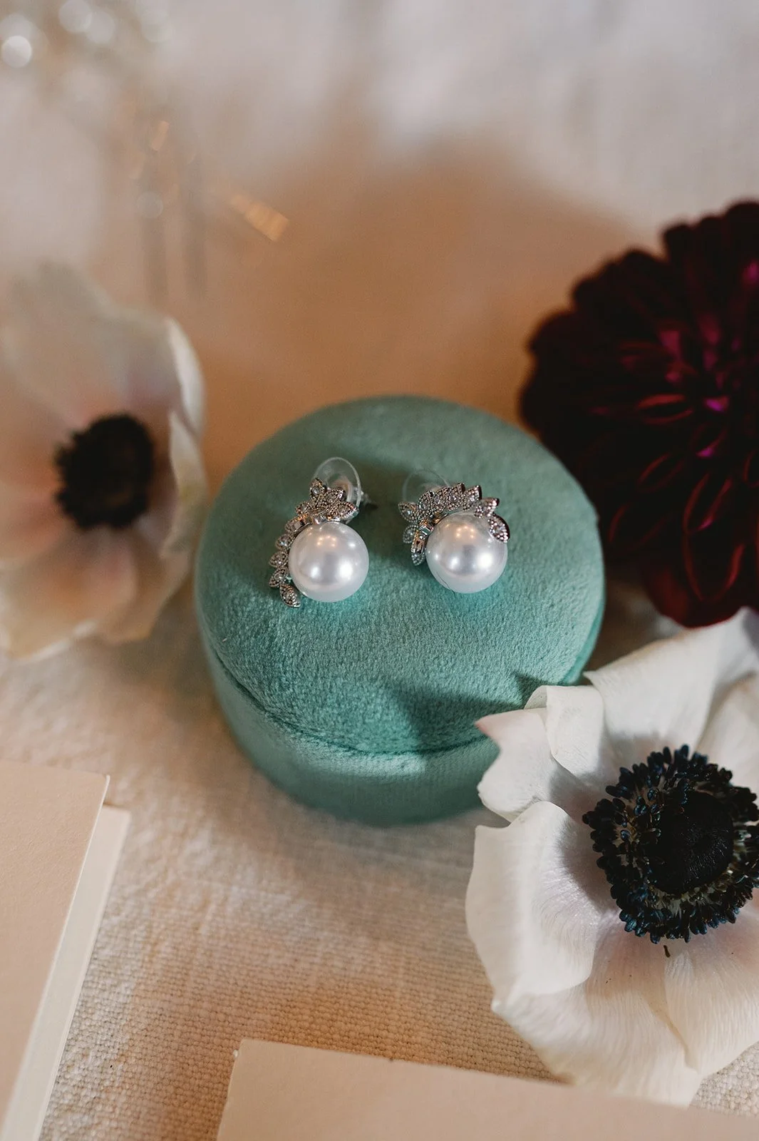 Pair of pearl earrings with rhinestone embellishments on a green jewelry display surrounded by white, black, and dark red flowers.