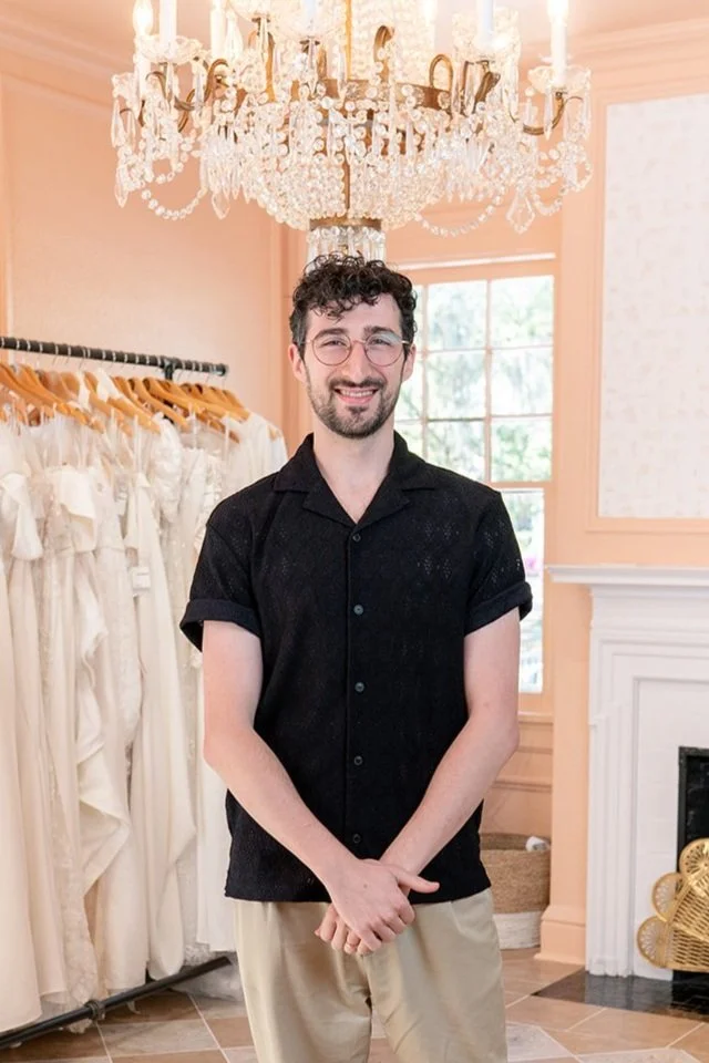 A man with glasses and curly hair smiling, standing inside a boutique with dresses on a rack behind him and a chandelier hanging above.