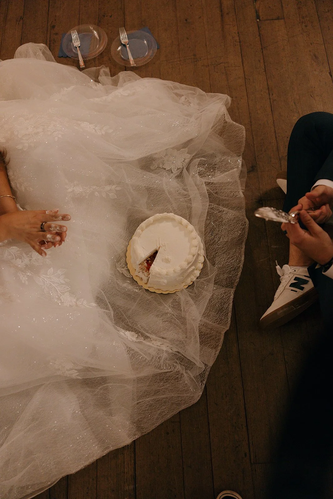Bride in a wedding dress and groom in a suit sitting on a wooden floor with a wedding cake and two empty plates in front of them.