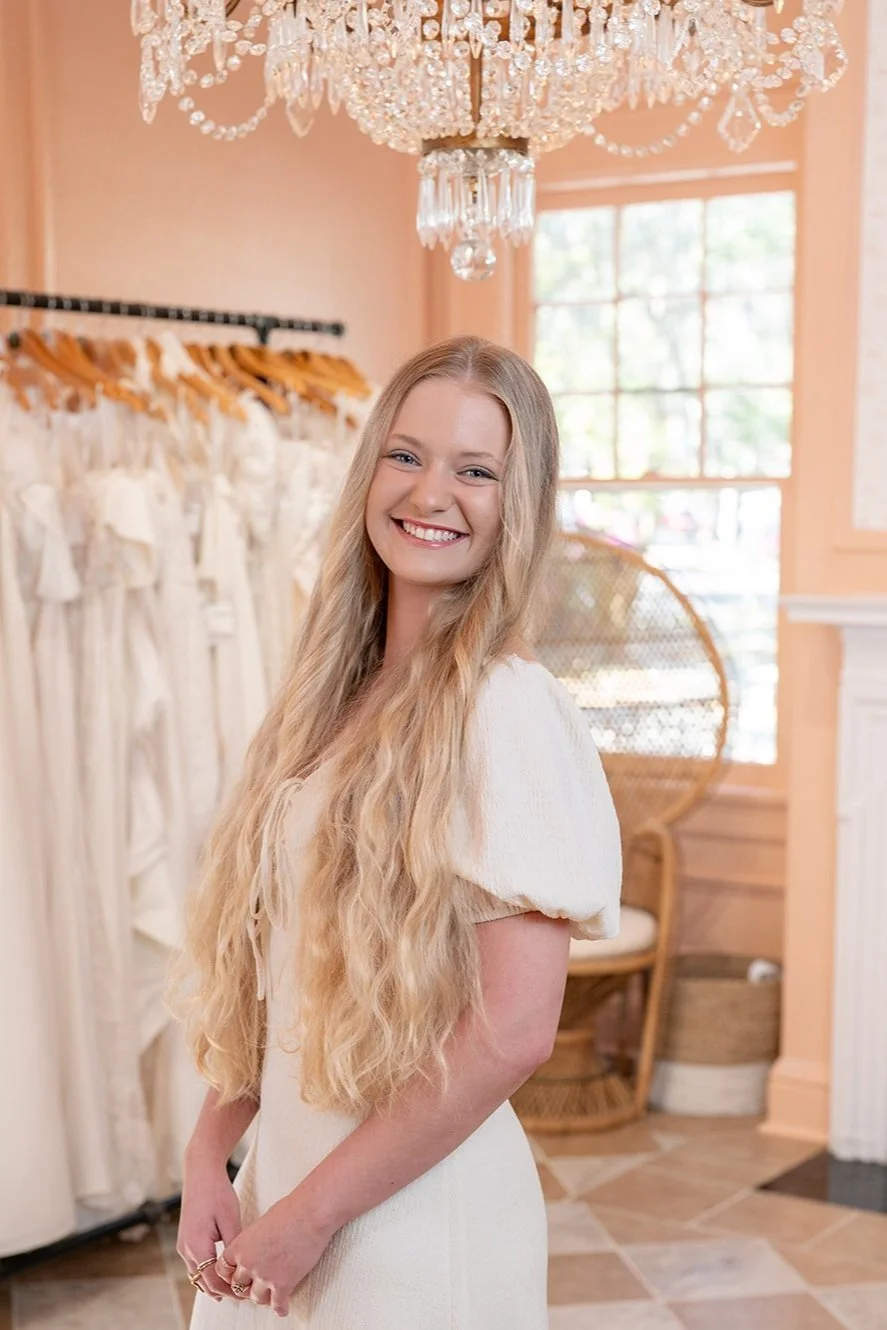 A young woman with long, wavy blonde hair smiling in a boutique with bridal dresses hanging behind her and a large window in the background.