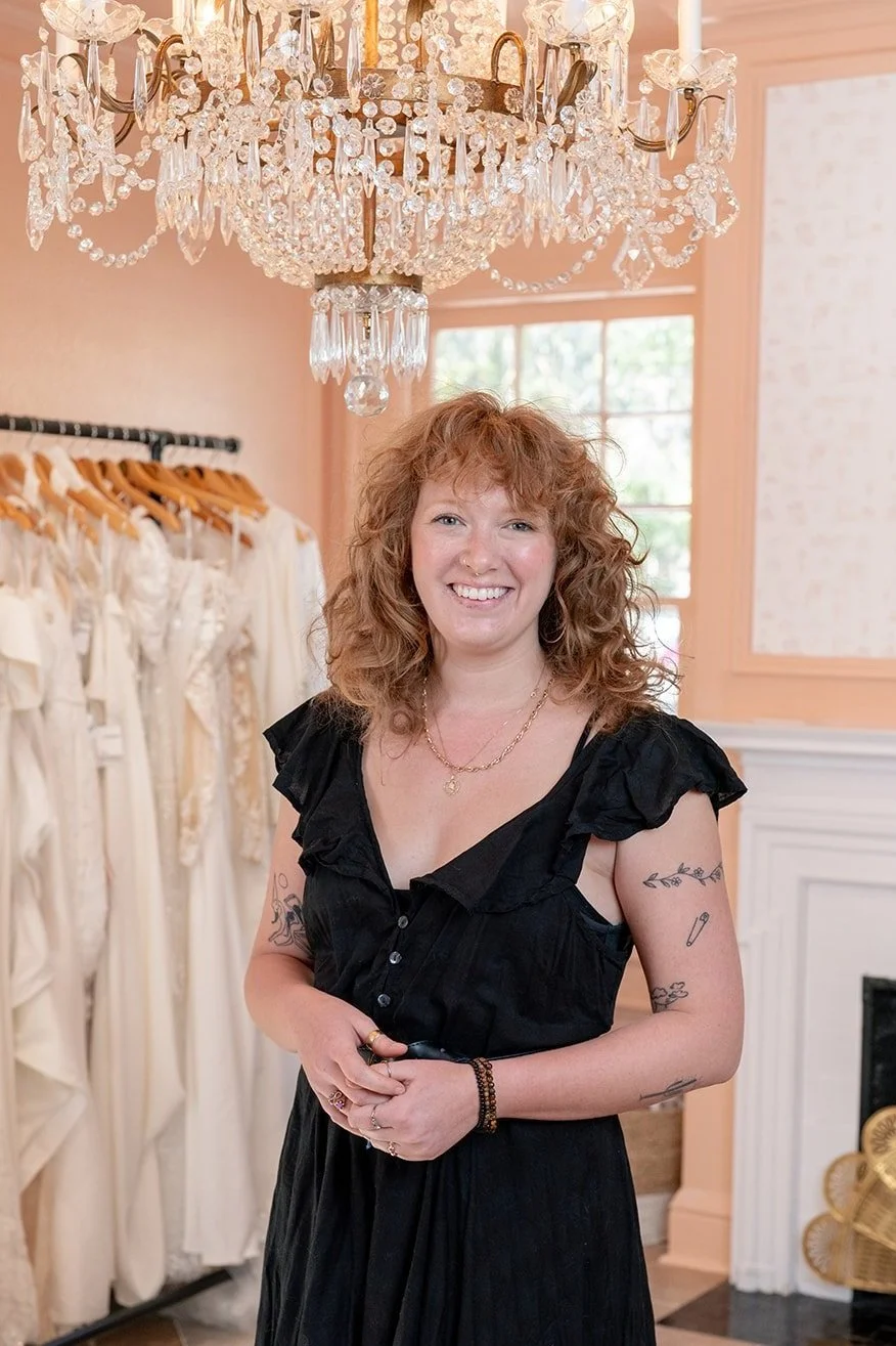 A woman with curly red hair and tattoos smiling in a boutique with ghosting dresses hanging on a rack behind her and a large crystal chandelier above.