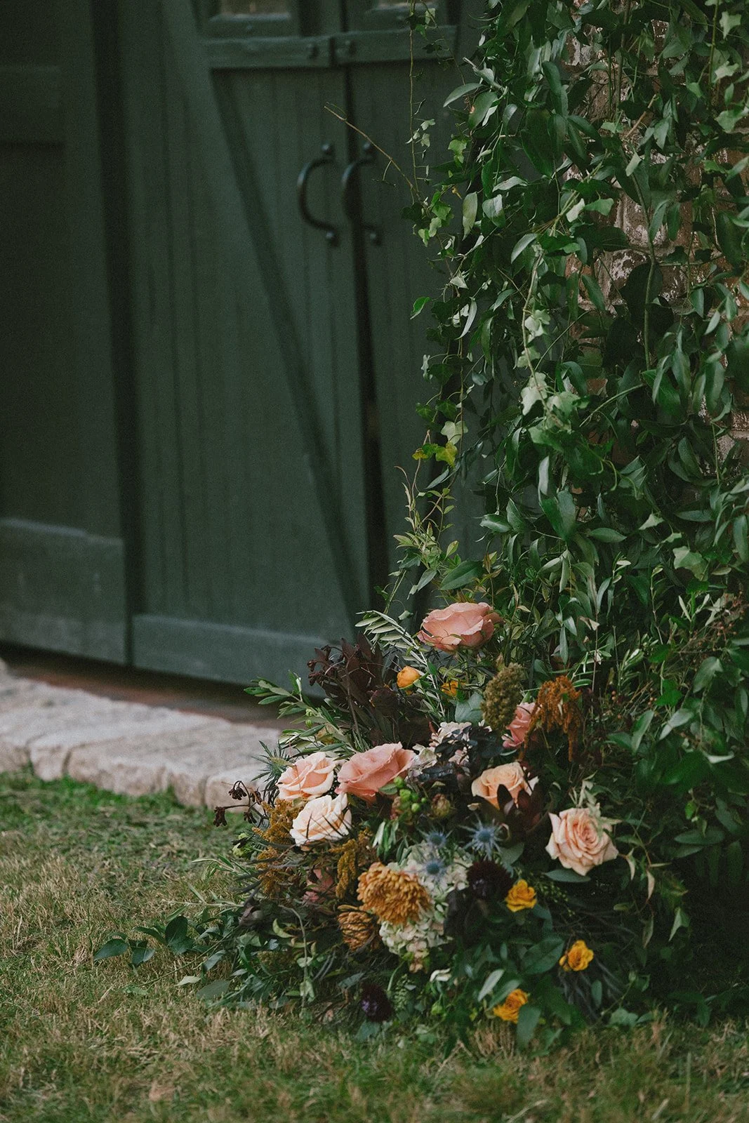 Organic wedding floral ground arrangement with peach roses, textured greenery and seasonal blooms beside historic green barn doors at Georgia State Railroad Museum wedding venue in Savannah Georgia.