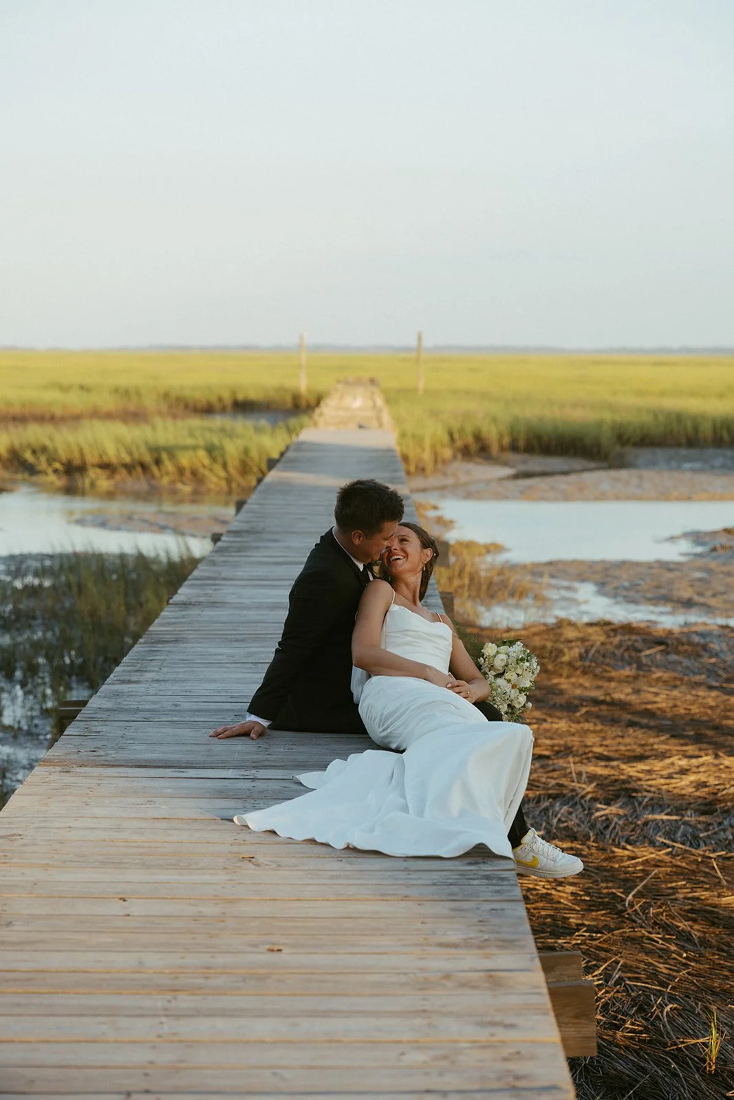 Bride and groom sitting on wooden dock overlooking marsh at sunset at Agapae Oaks wedding venue on St. Helena Island South Carolina Lowcountry destination wedding.