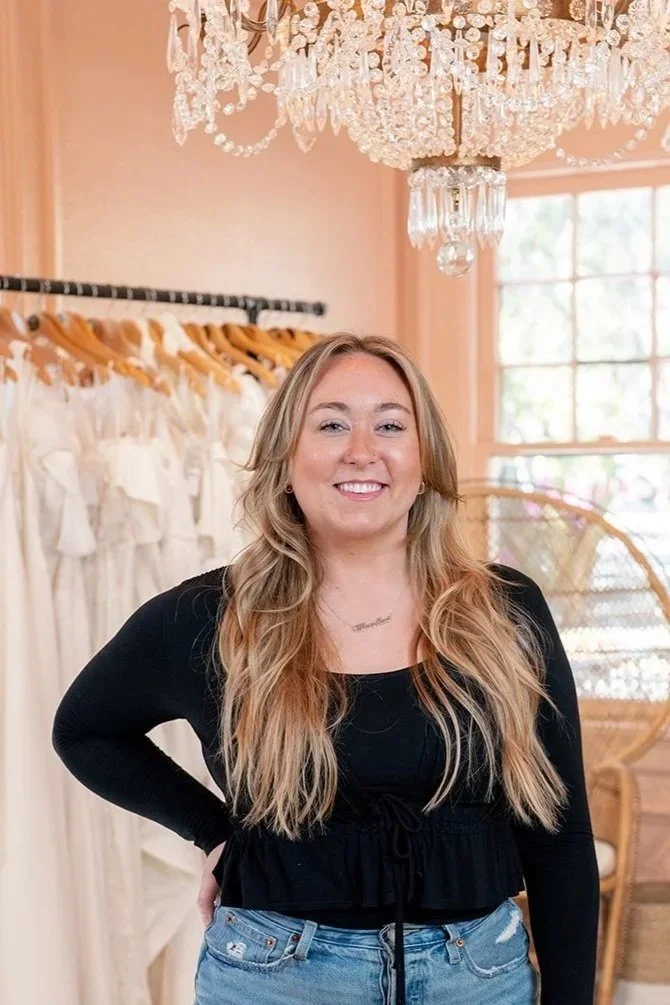A smiling woman with long blonde hair standing in a boutique or dressing room, surrounded by white dresses hanging on a black rack, with a large chandelier hanging above and a window in the background.