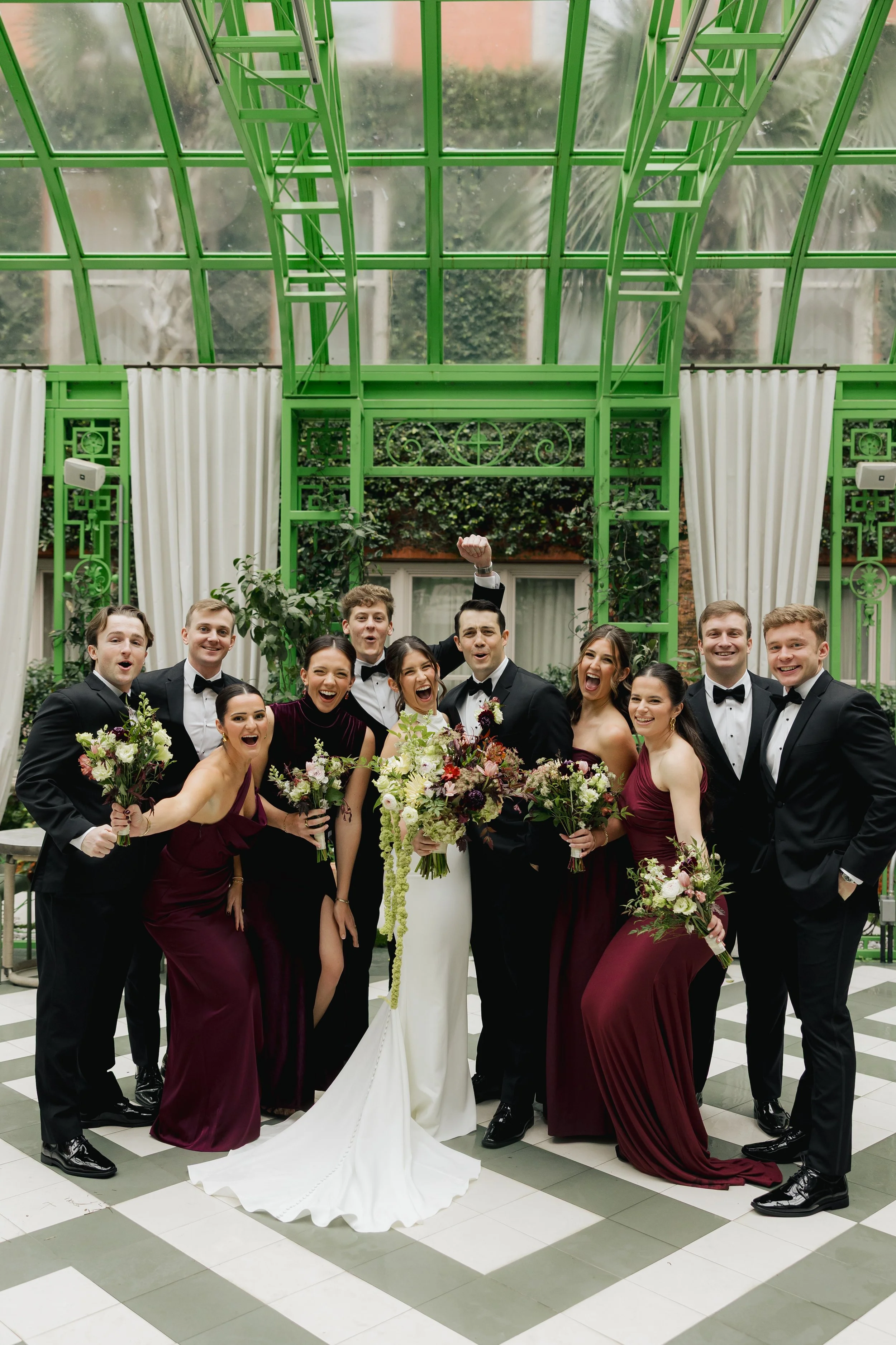 Wedding party celebrating beneath the green glass conservatory at Hotel Bardo Savannah, featuring burgundy bridesmaid dresses, classic tuxedos, and garden-style florals by Ivory & Beau.