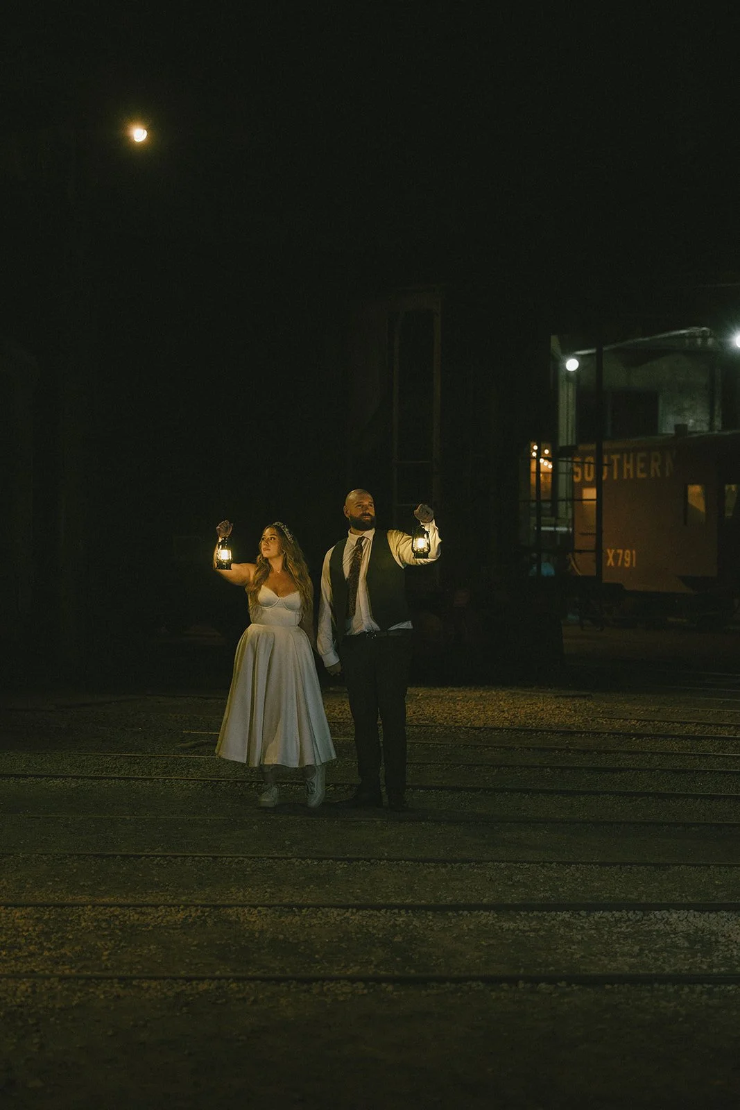 Bride and groom walking with lanterns at night on railroad tracks during Georgia State Railroad Museum wedding in Savannah Georgia destination wedding venue.