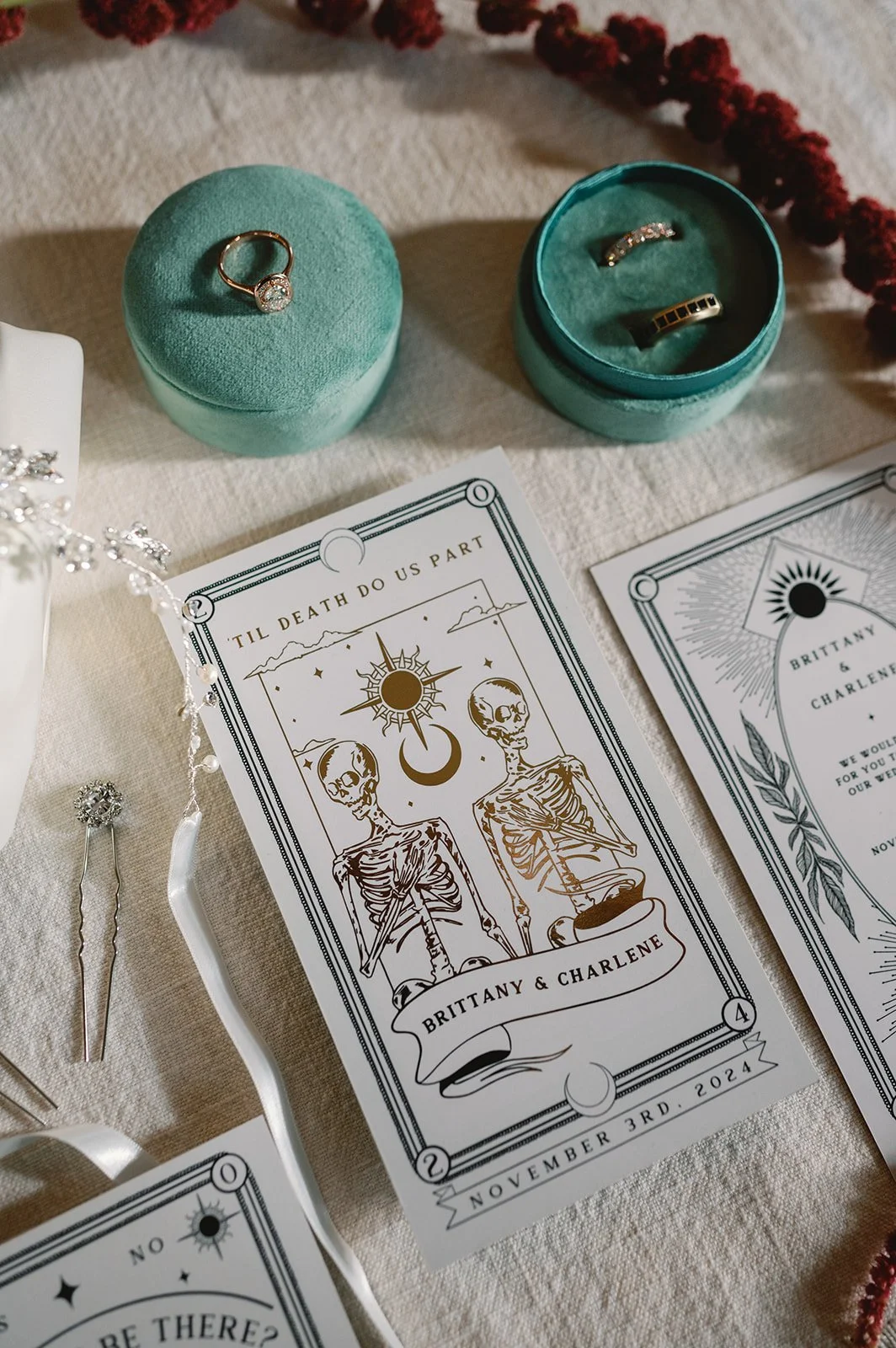 Wedding invitation for Brittany and Charlene dated November 3rd, 2024, featuring skeletons and celestial symbols, with rings in teal boxes and a silver hairpin with a flower design.