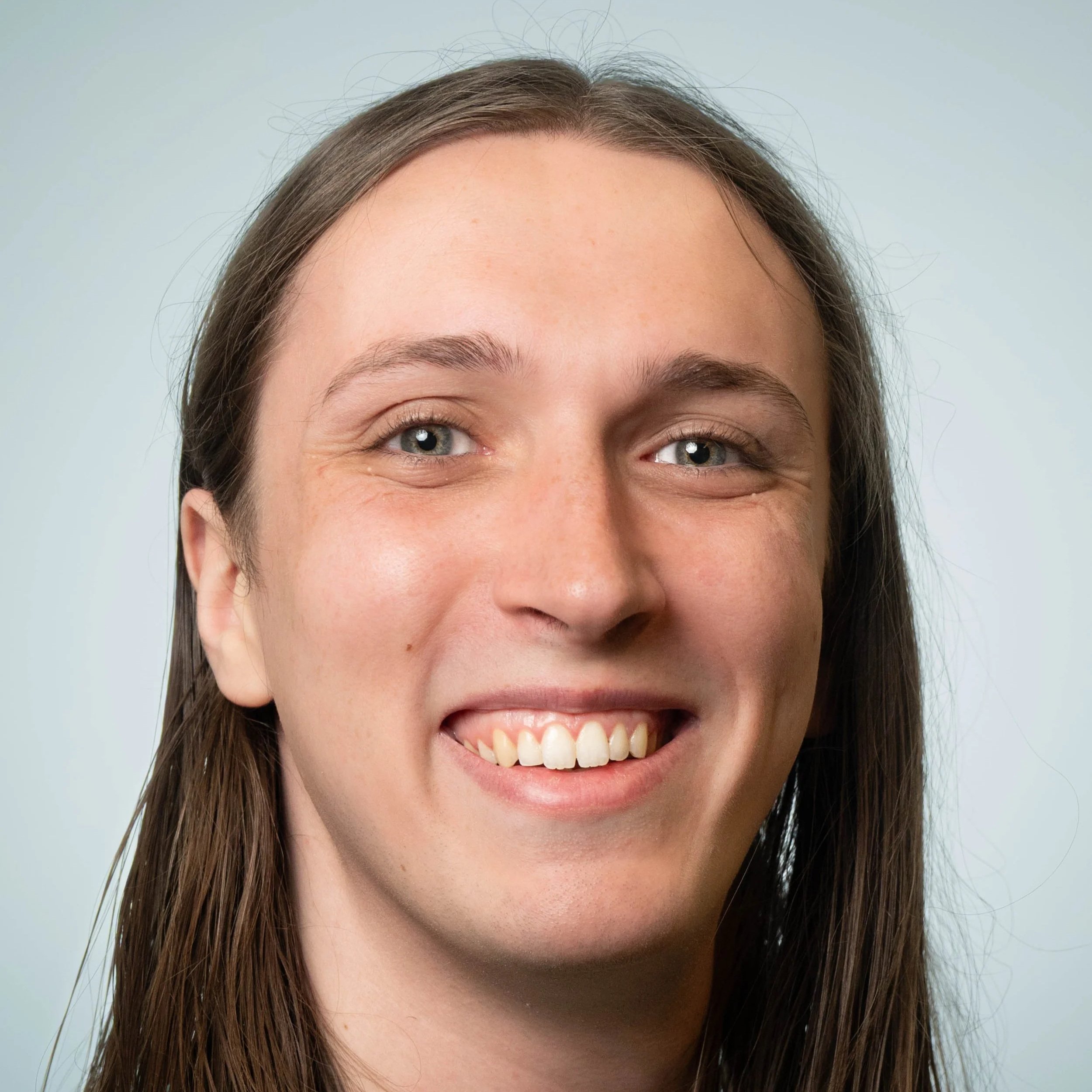 Portrait of a young woman with long brown hair, blue eyes, and a bright smile, wearing a turquoise shirt, standing in front of a plain light-colored wall.