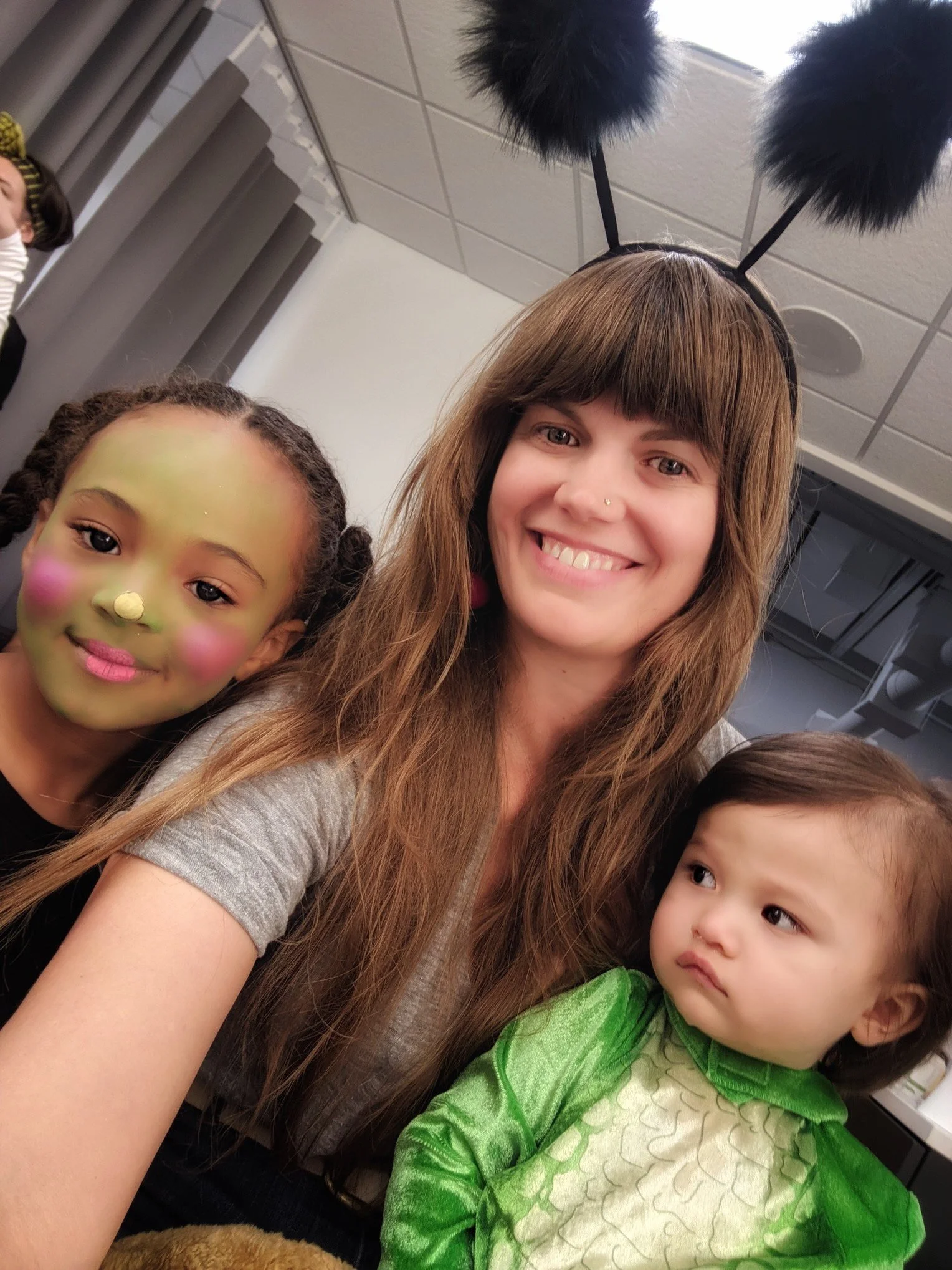 Two young girls and a woman taking a selfie in costumes, one girl has face paint resembling a green dragon, and the other girl is dressed as a dragon, in what appears to be a classroom or indoor setting.