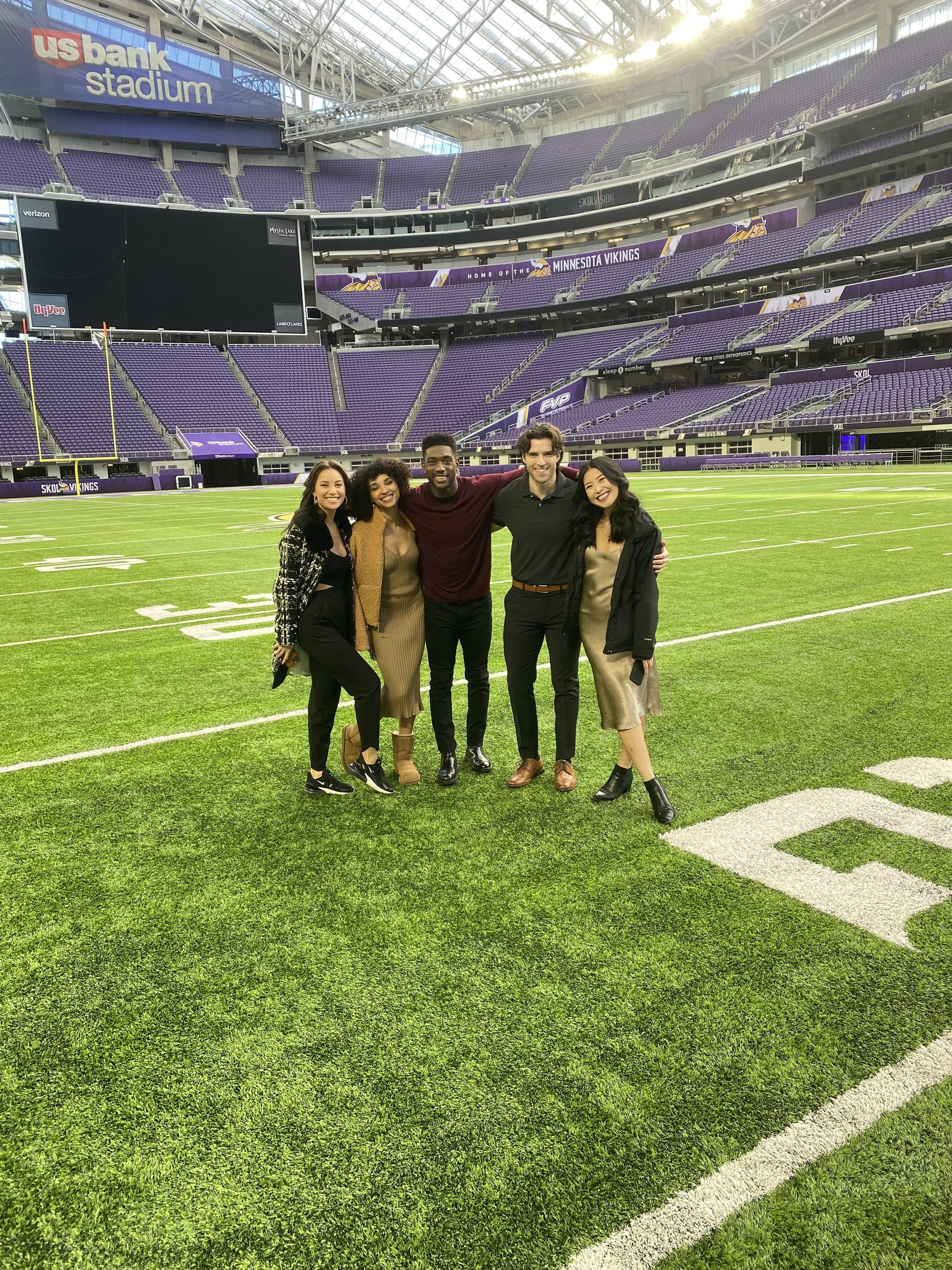 Five people standing on a football field inside a stadium, smiling, with empty purple seats and a large screen in the background that reads "US Bank Stadium" and "Home of the Minnesota Vikings."