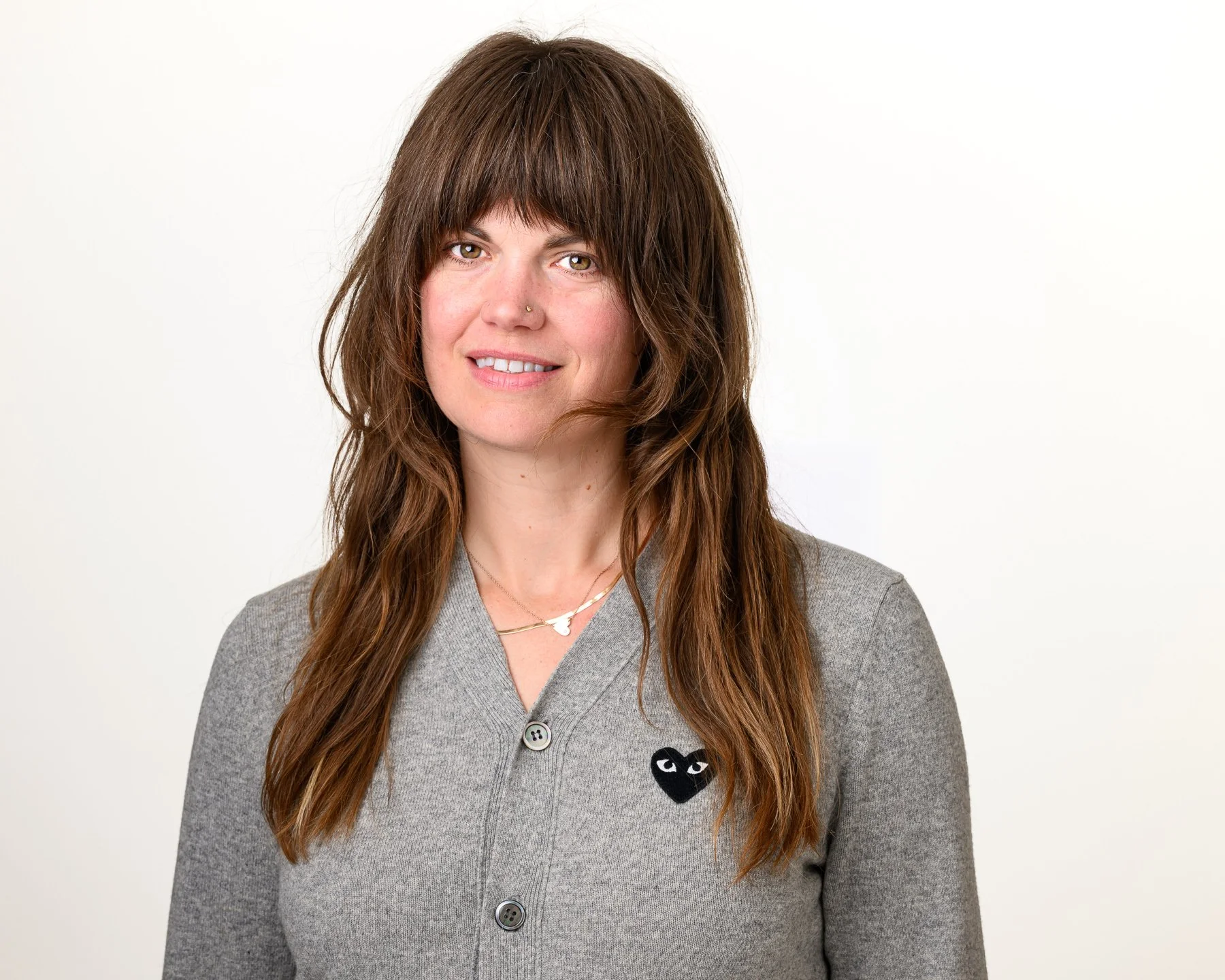 A woman with long, wavy brown hair and hazel eyes, wearing a grey cardigan with a pokémon logo on it, gold jewelry, and a nose ring, standing against a plain white background.