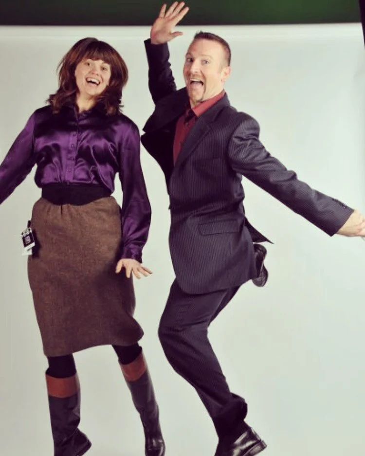 Man and woman jumping and smiling, dressed in business attire, against a plain background.