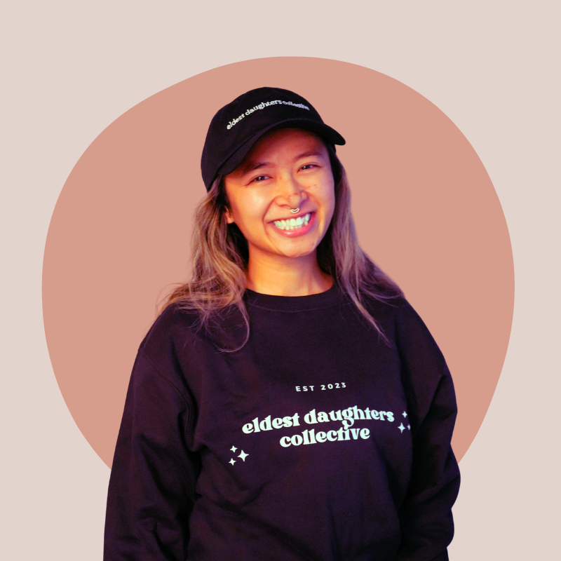 A young woman smiling, wearing a black cap and sweatshirt with the words "eldest daughters collective," against a beige background with a pinkish circle.