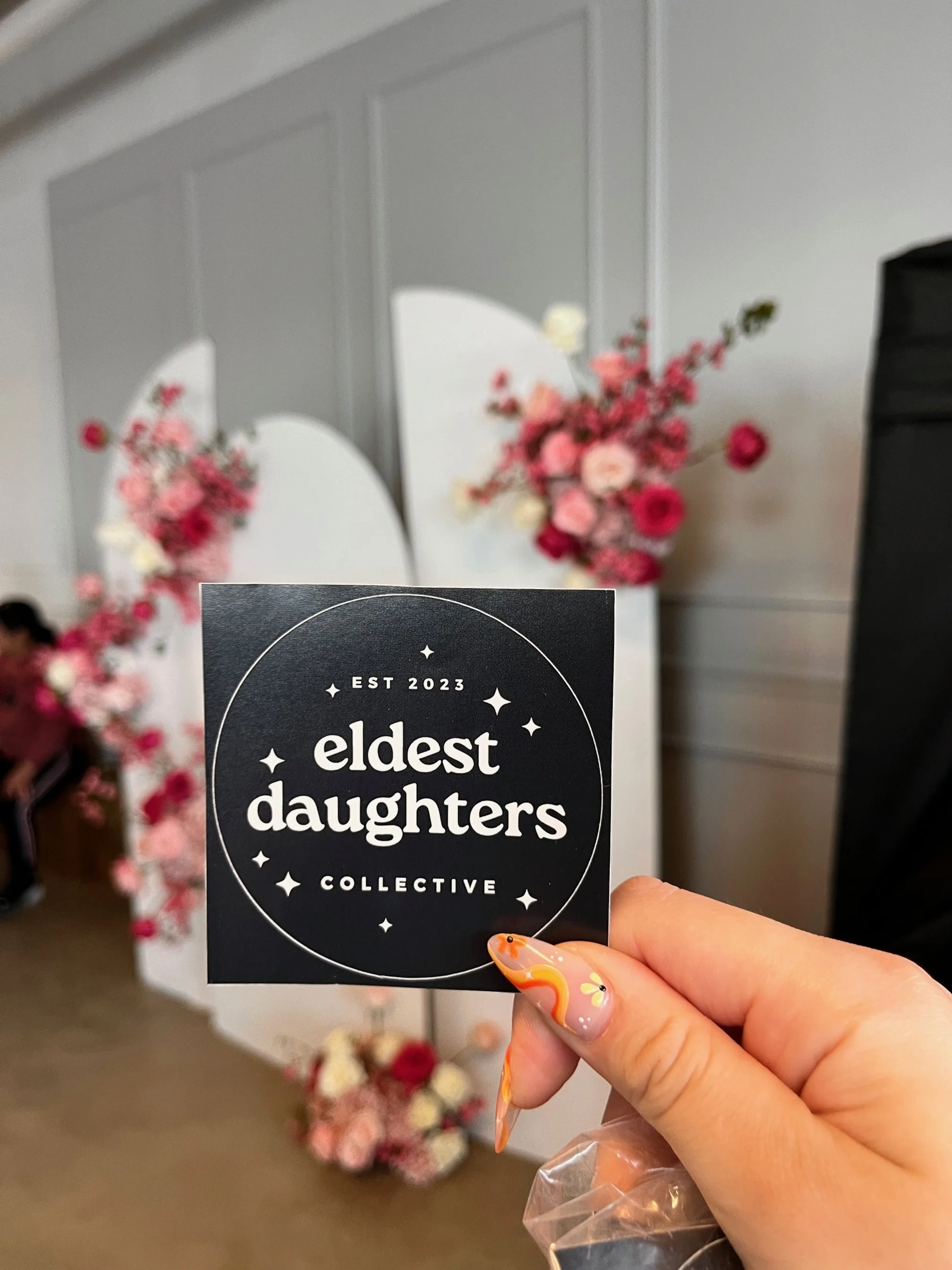 A hand holding a black circular sticker that reads 'eldest daughters collective' with 'EST 2023' at the top, in front of a background with floral arrangements and a white mirror.