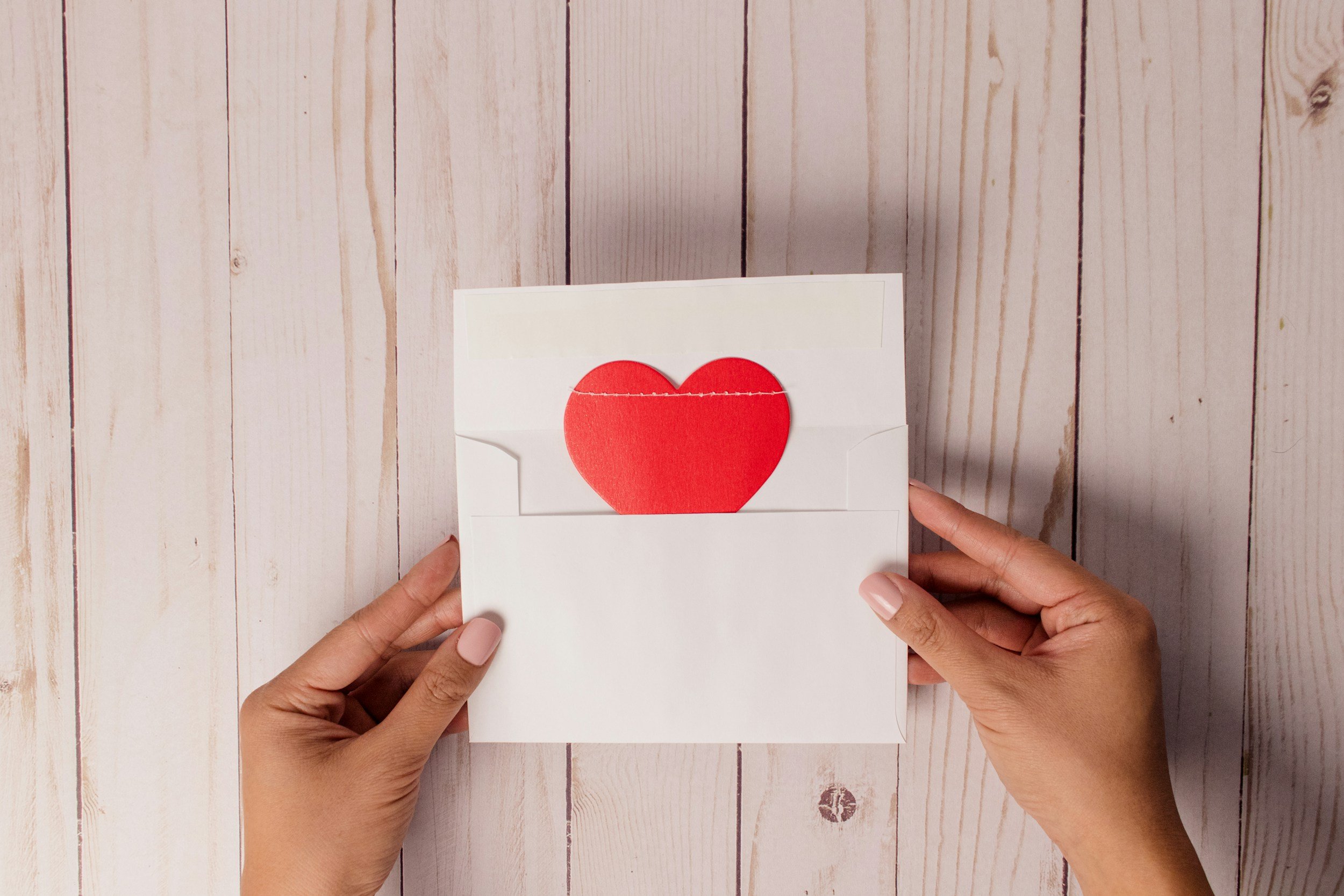 A person holding a white envelope with a red paper heart inside, on a light wooden background.