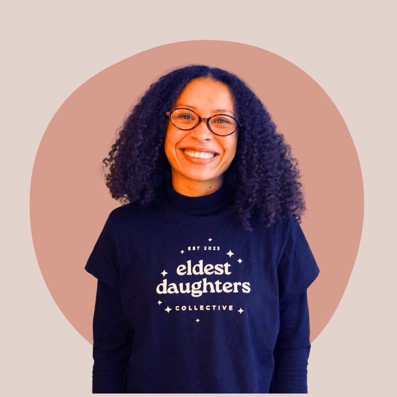 Woman with curly dark hair and glasses smiling, wearing a black t-shirt that says "eldest daughters collective" with small stars and "Est 2023" lettering, standing in front of a beige and pink background.