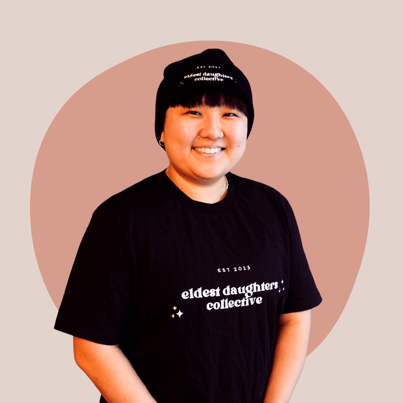 A smiling young woman wearing a black beanie and black T-shirt with white text that says 'eldest daughters collective' and a beige background with a large pinkish circle behind her.