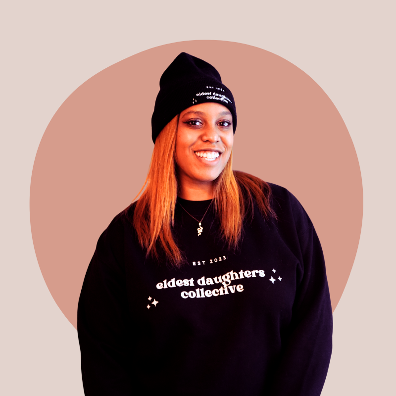 A smiling woman with shoulder-length red hair, wearing a black beanie and a black sweatshirt with white text that says "eldest daughters collective" and a necklace with a small pendant. Background features a circular beige and light brown gradient.