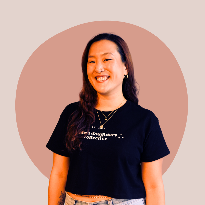 A woman with long dark hair smiling, wearing a black t-shirt with white text, layered necklaces, and earrings, standing in front of a pinkish-beige background with a circle design.