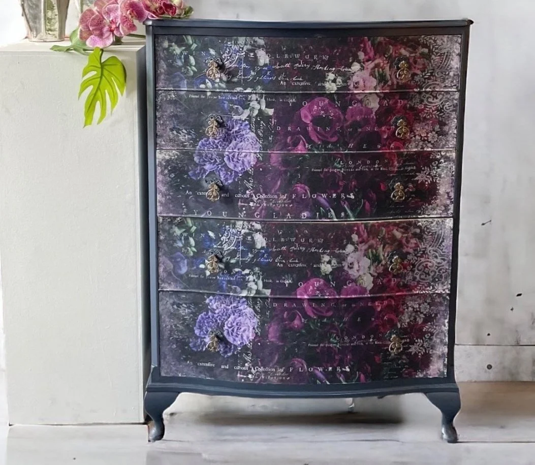 Black dresser with floral design and butterflies, featuring drawers with ornate handles, placed against a white wall with a white surface on top, decorated with pink flowers and green leaves.