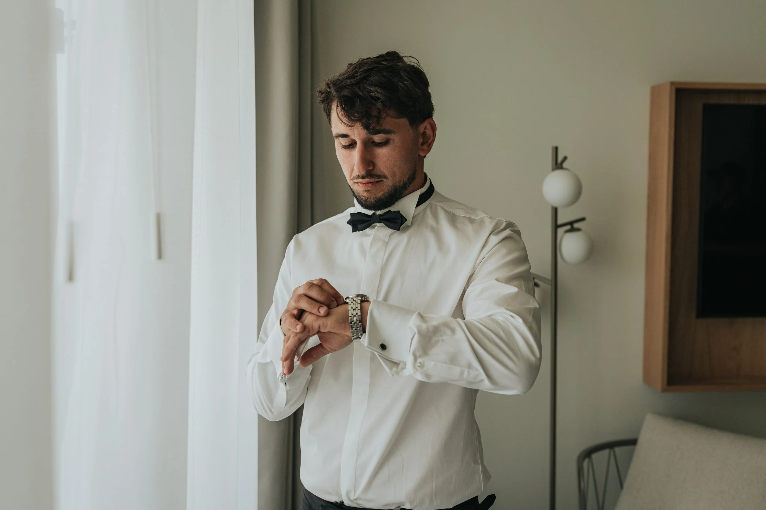 A young man with dark hair and a beard, dressed in a white shirt and black bow tie, is looking at his wristwatch in a well-lit room near a window with curtains.
