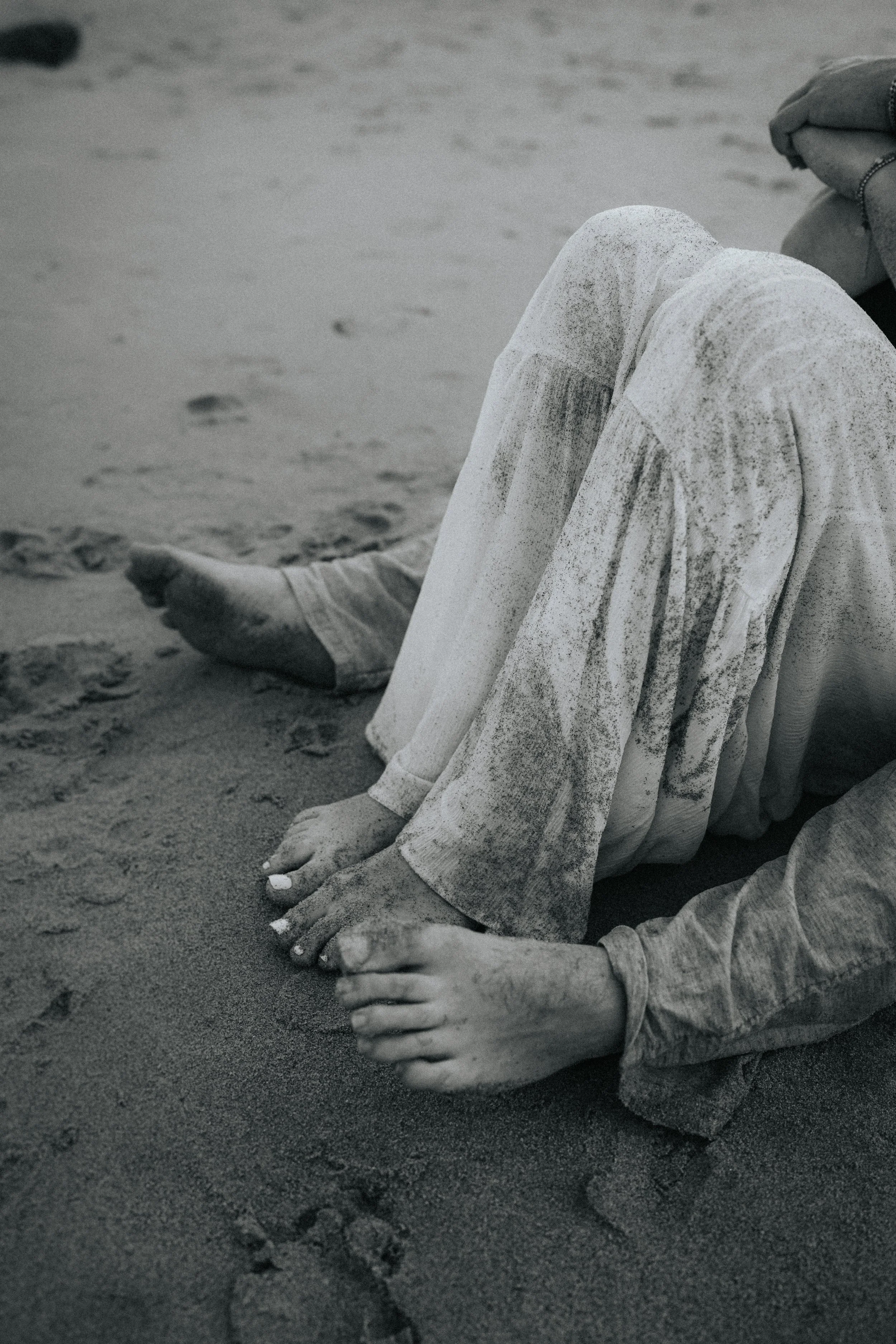 Close-up of a person sitting on the sand at the beach. The person is wearing light-colored jeans and a long-sleeve shirt, with their hands resting on the sand and legs pulled up. The image is in black and white.