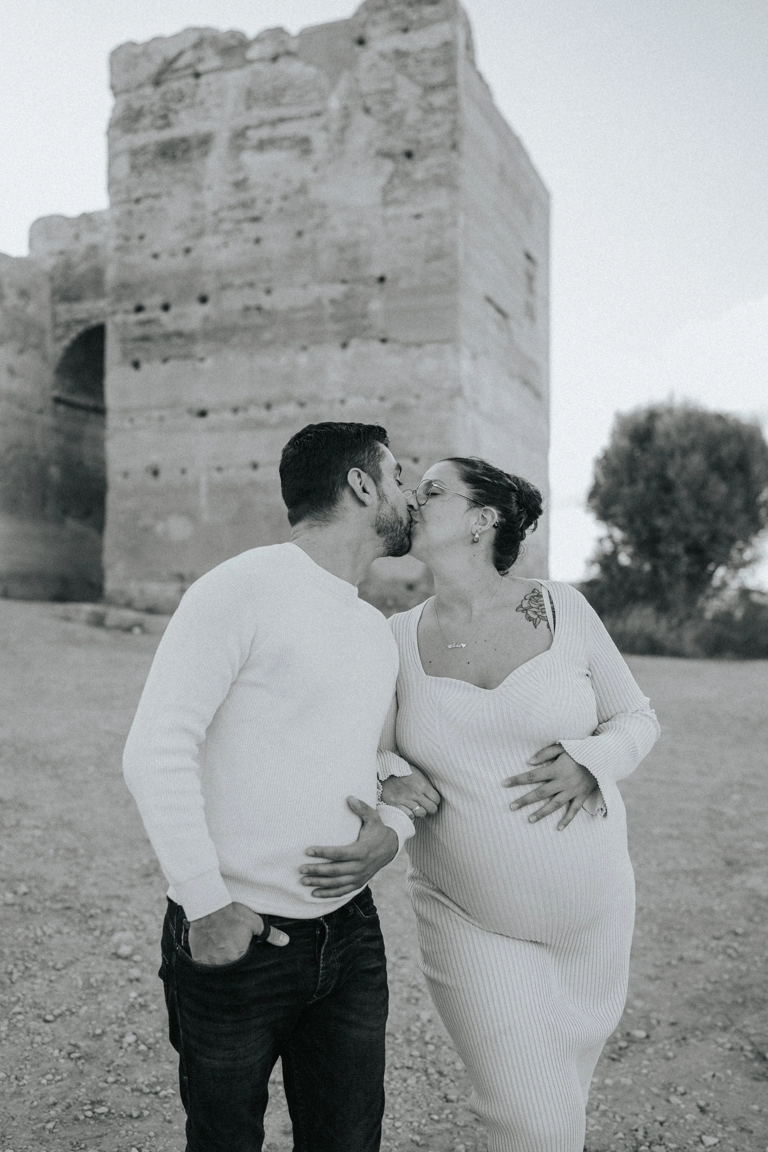 A black-and-white photo of a couple kissing outdoors in front of an ancient stone structure with arches and a large tree in the background.