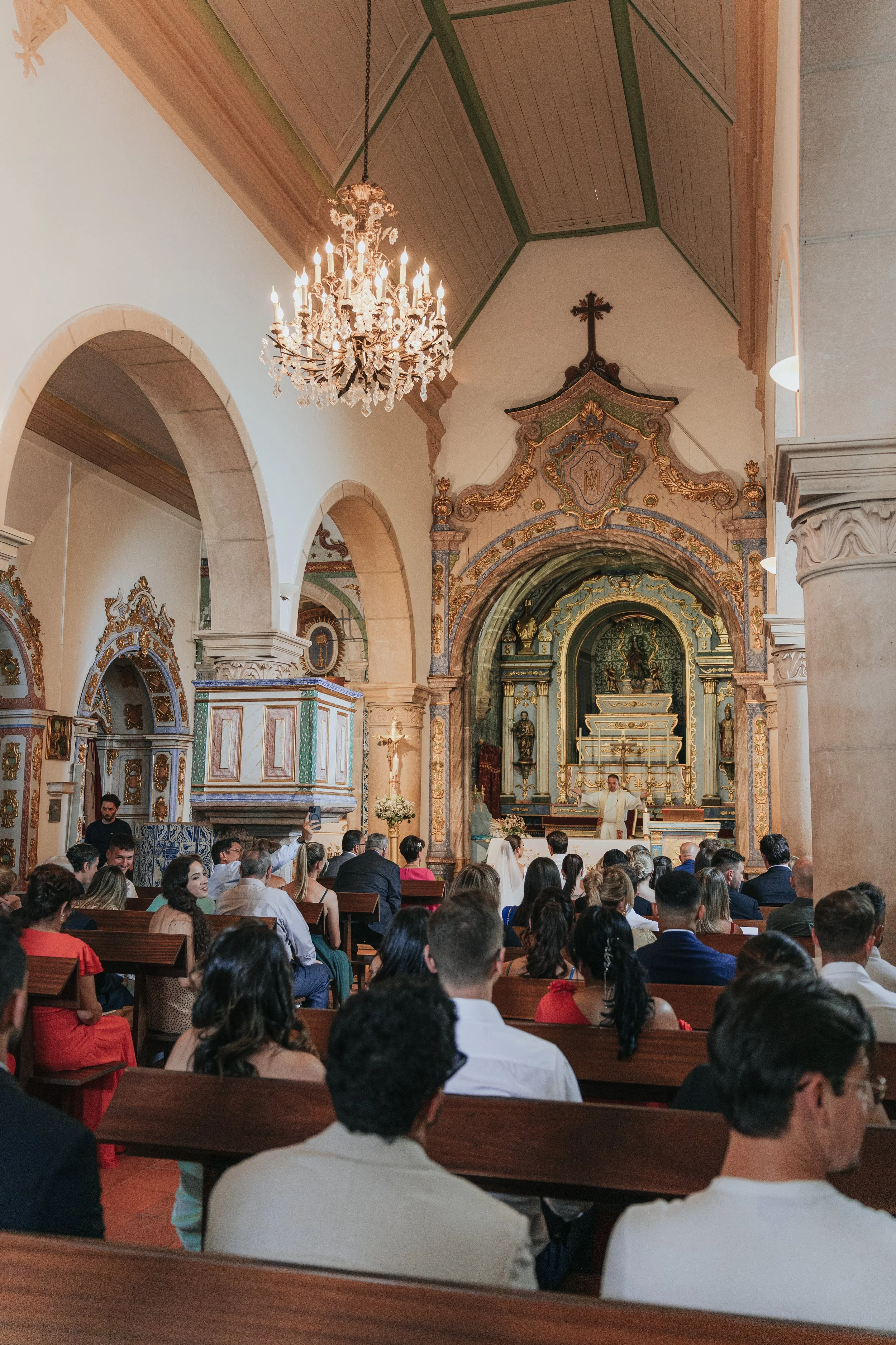 Inside a church during a wedding ceremony with many seated guests, a priest officiating at the altar, ornate decorations, wood ceiling, chandelier, and religious symbols.
