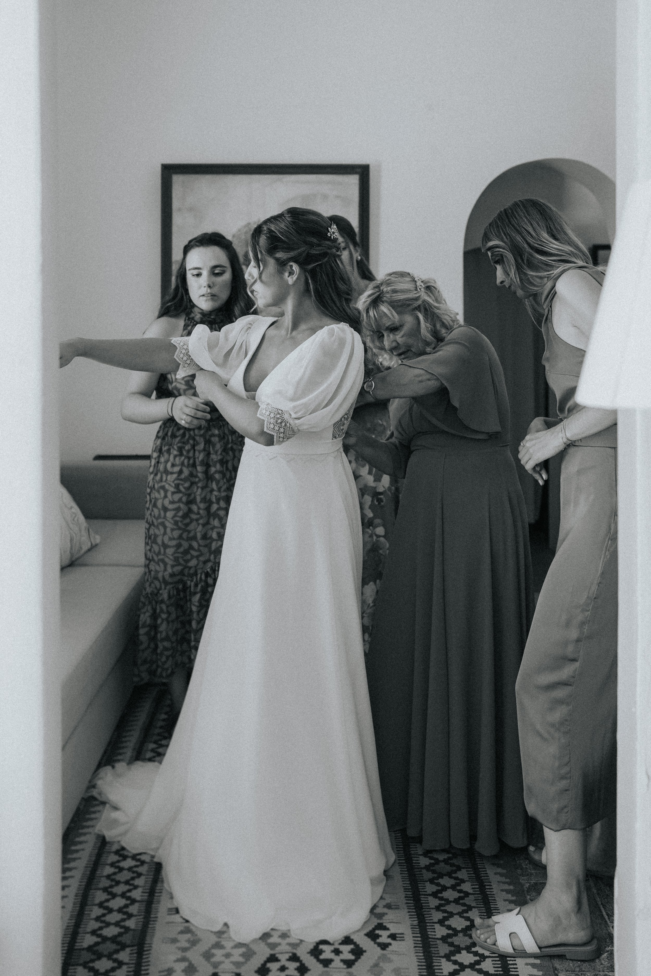 A group of women, likely preparing for a wedding, in a room with a patterned floor, some adjusting a wedding dress and others helping.